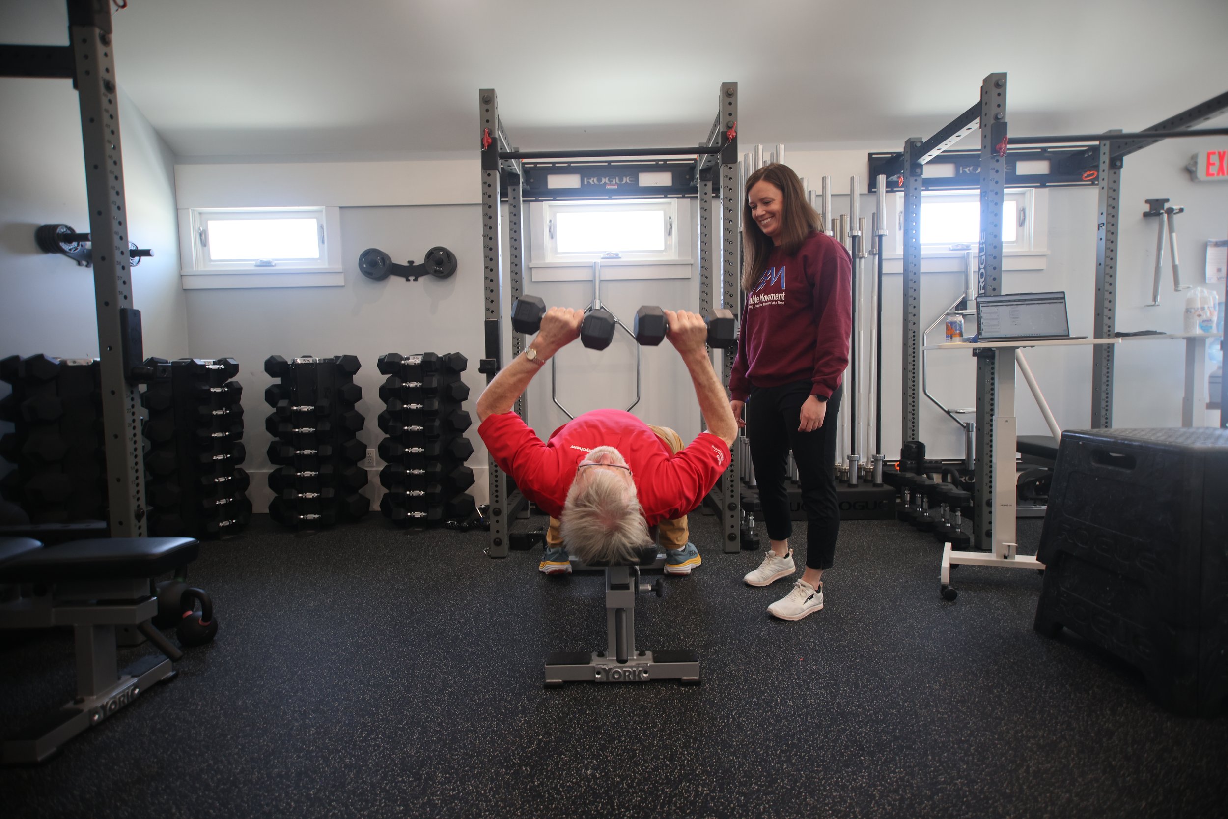 An elderly woman with gray hair in a red shirt is doing a bench press exercise with dumbbells in a gym, while a young woman observing and smiling stands beside her.