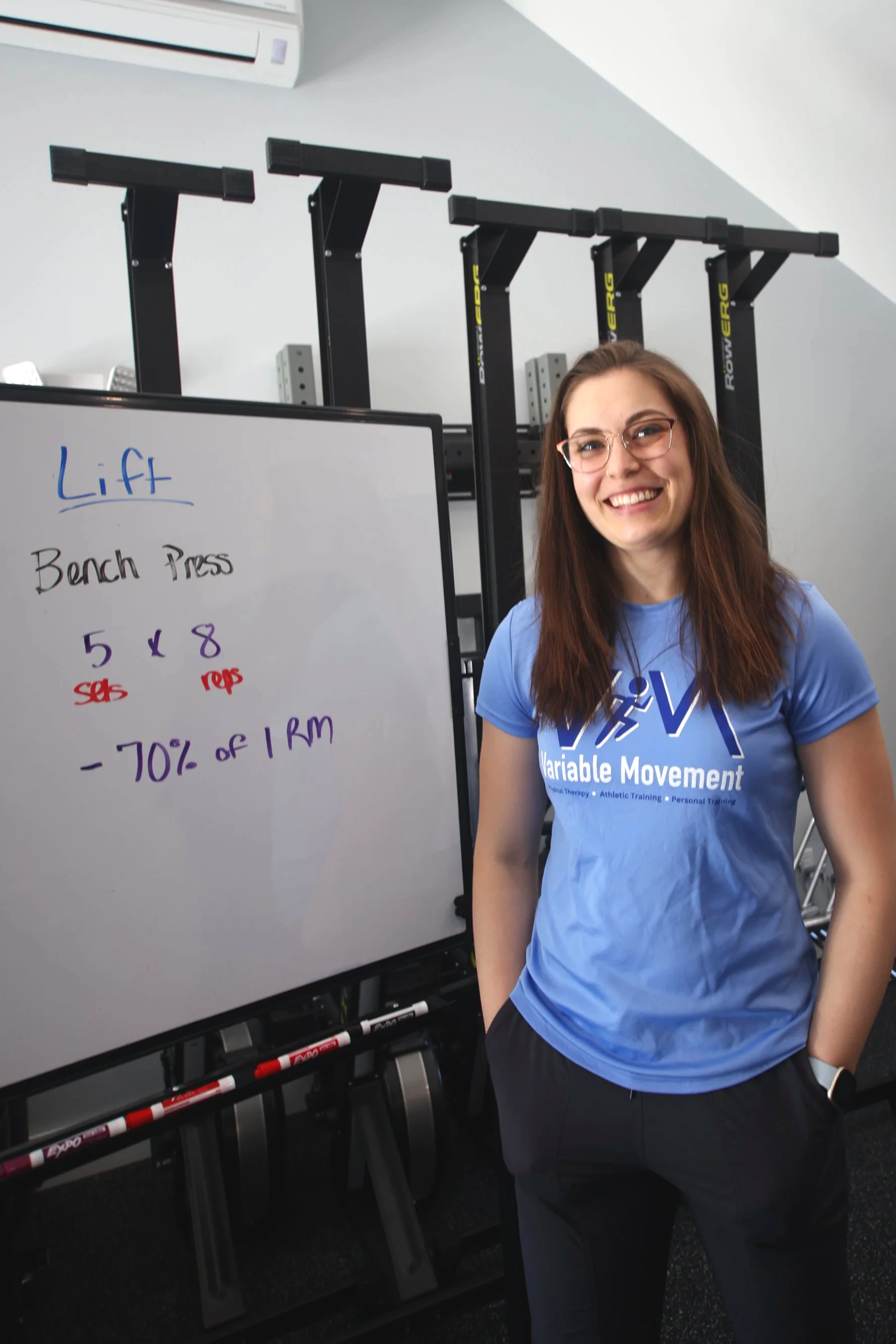 A young woman smiling, wearing glasses and a blue shirt that says 'Variable Movement' is standing in a gym or rehabilitation facility.