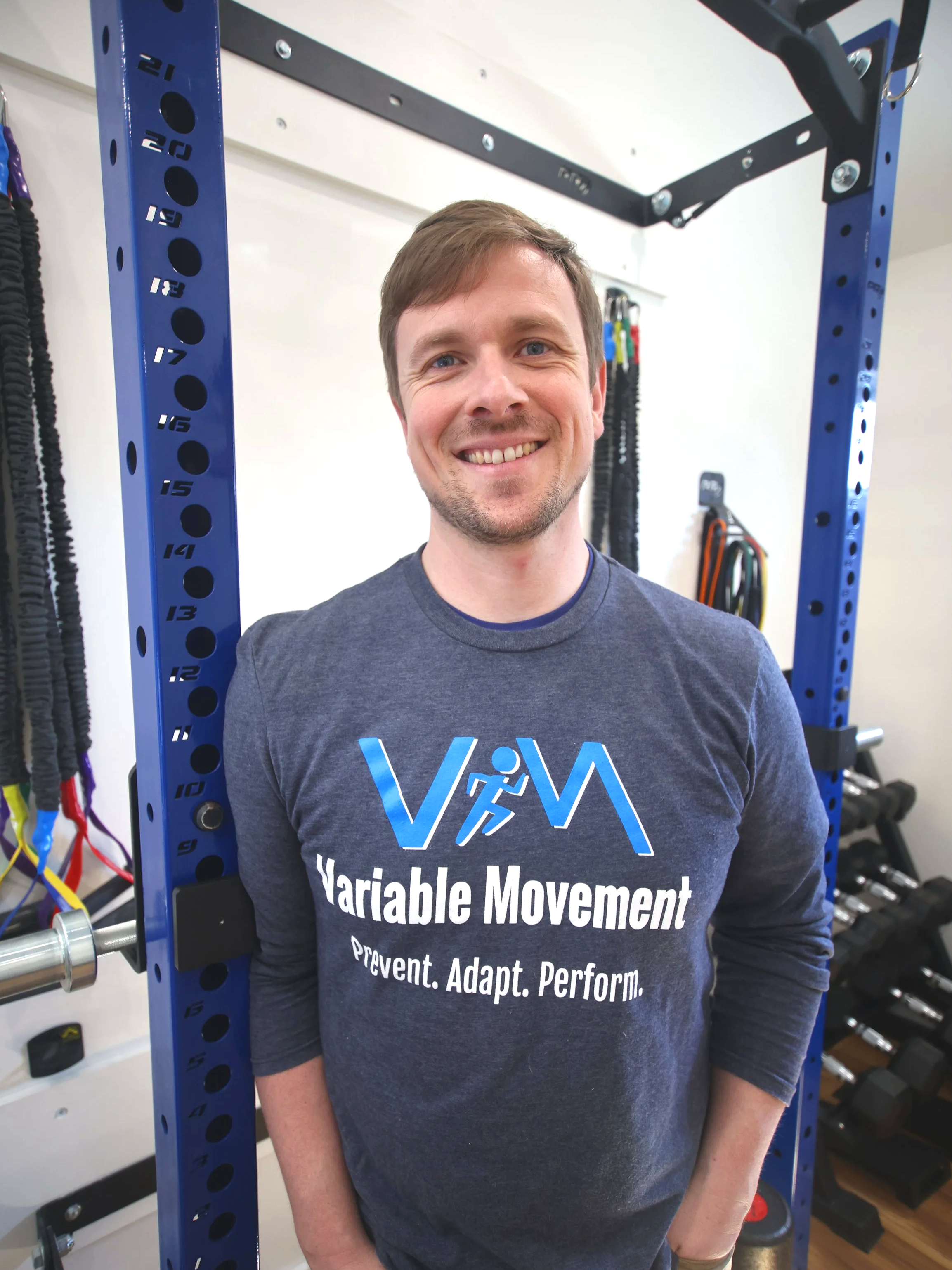 A smiling man standing in a gym next to a blue power rack, wearing a dark gray shirt with 'VAM' and 'Variable Movement' printed on it.