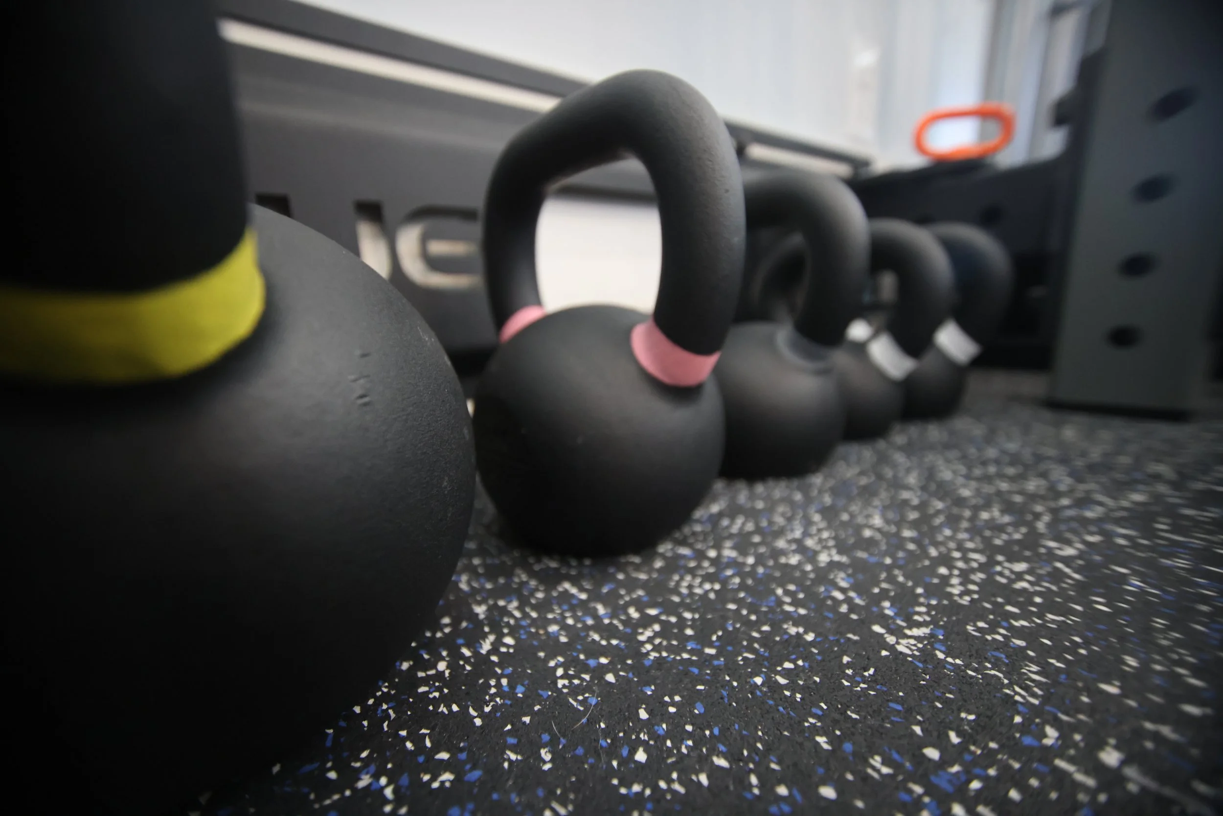 Black kettlebells with pink and white markings lined up on a speckled gym floor near exercise equipment.