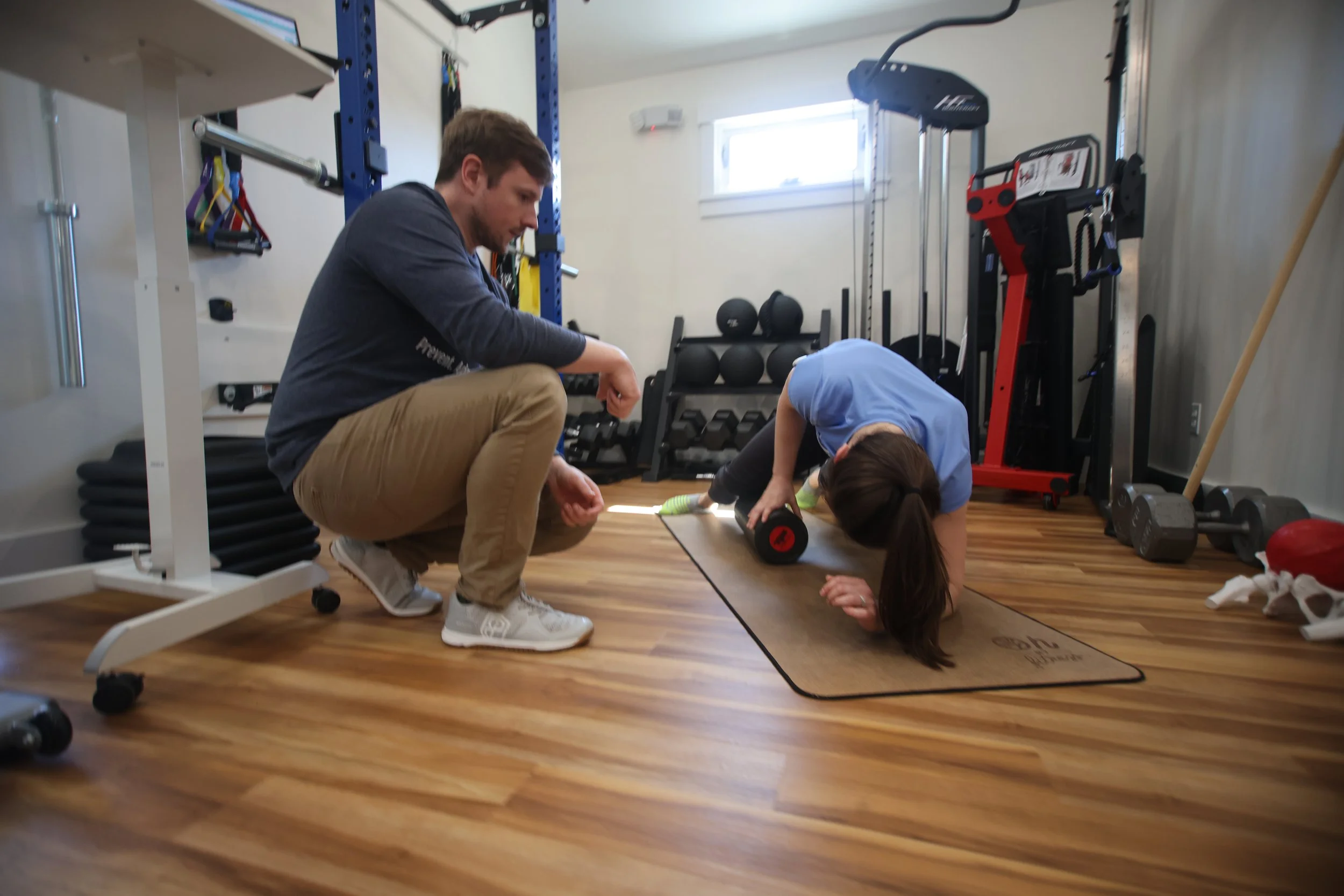 A woman is performing an exercise with a foam roller on a workout mat in a home gym, while a man kneels beside her providing guidance.