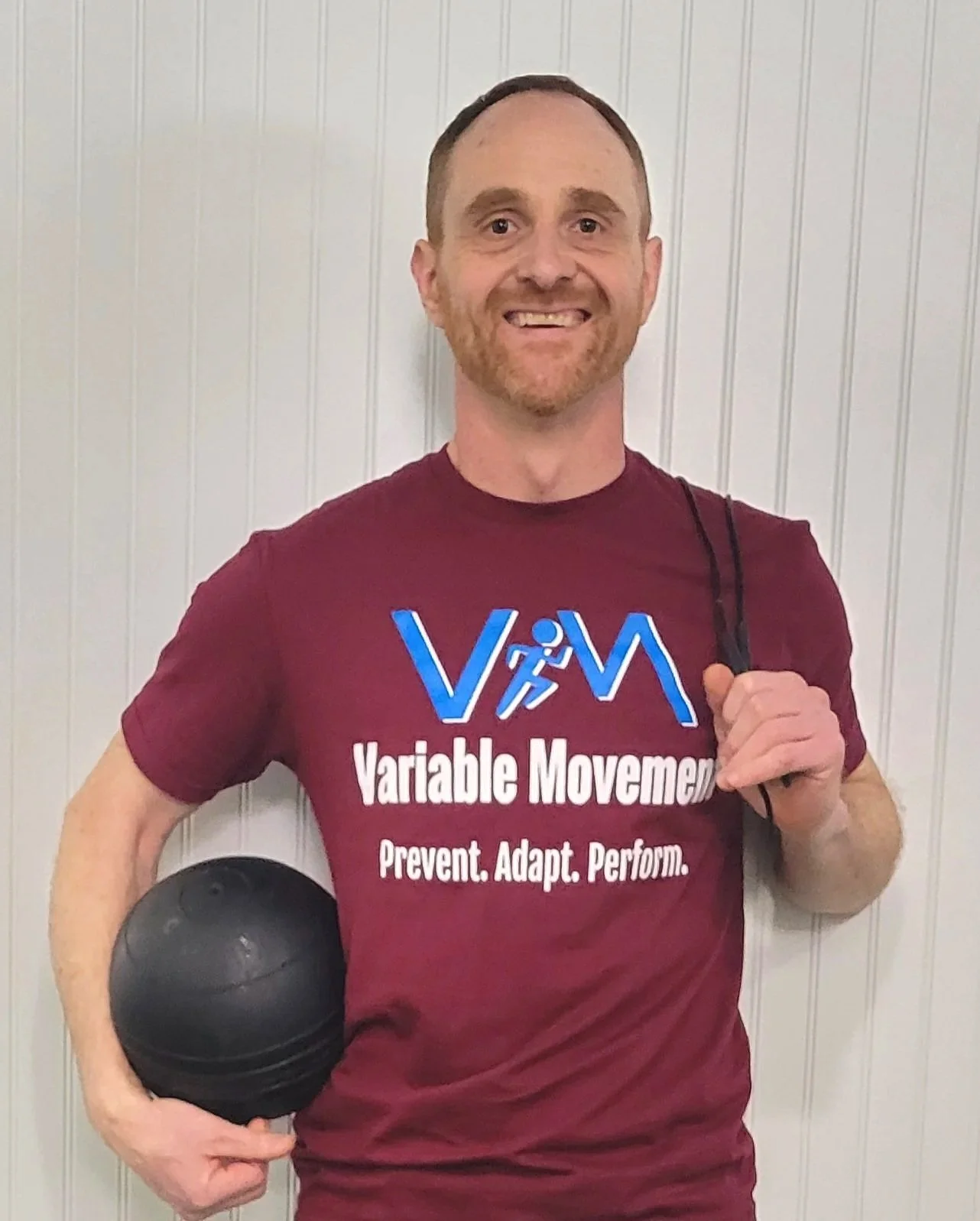 Blue Hill personal trainer wearing a maroon T-shirt with the text 'Variable Movement' and a logo, against a light-colored, paneled wall.