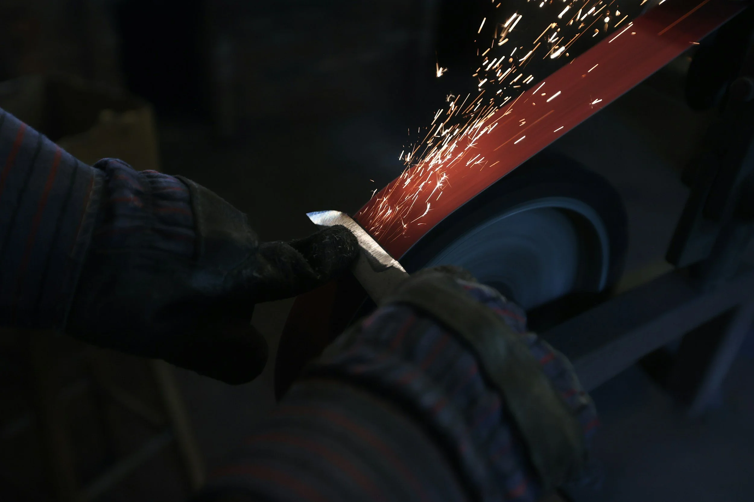 A person wearing gloves uses a grinder to smooth the edge of a red metal piece, creating sparks.