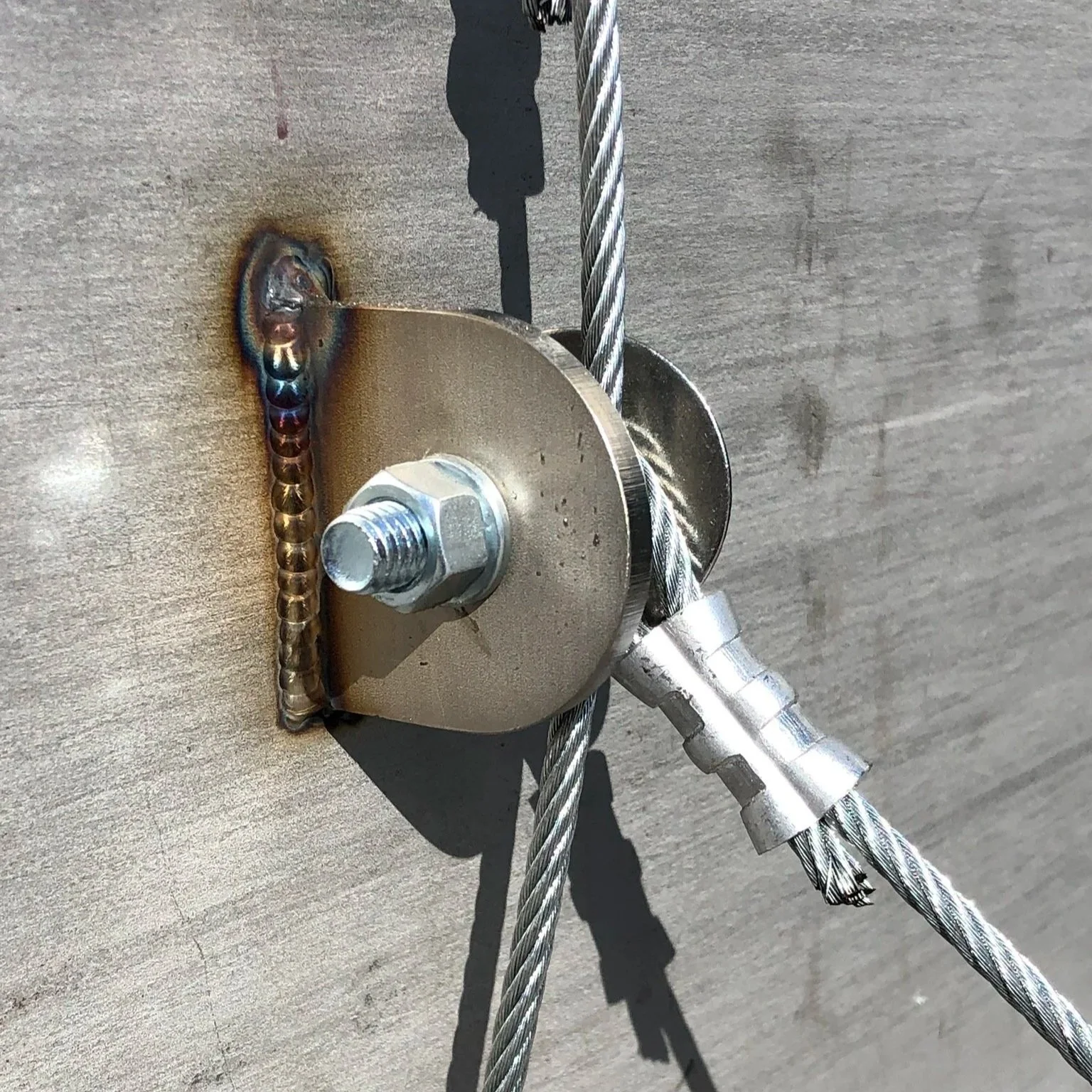 Close-up of a metal wire rope attached to a wall with a metal plate, bolt, and nut, with welding around the plate's edges.