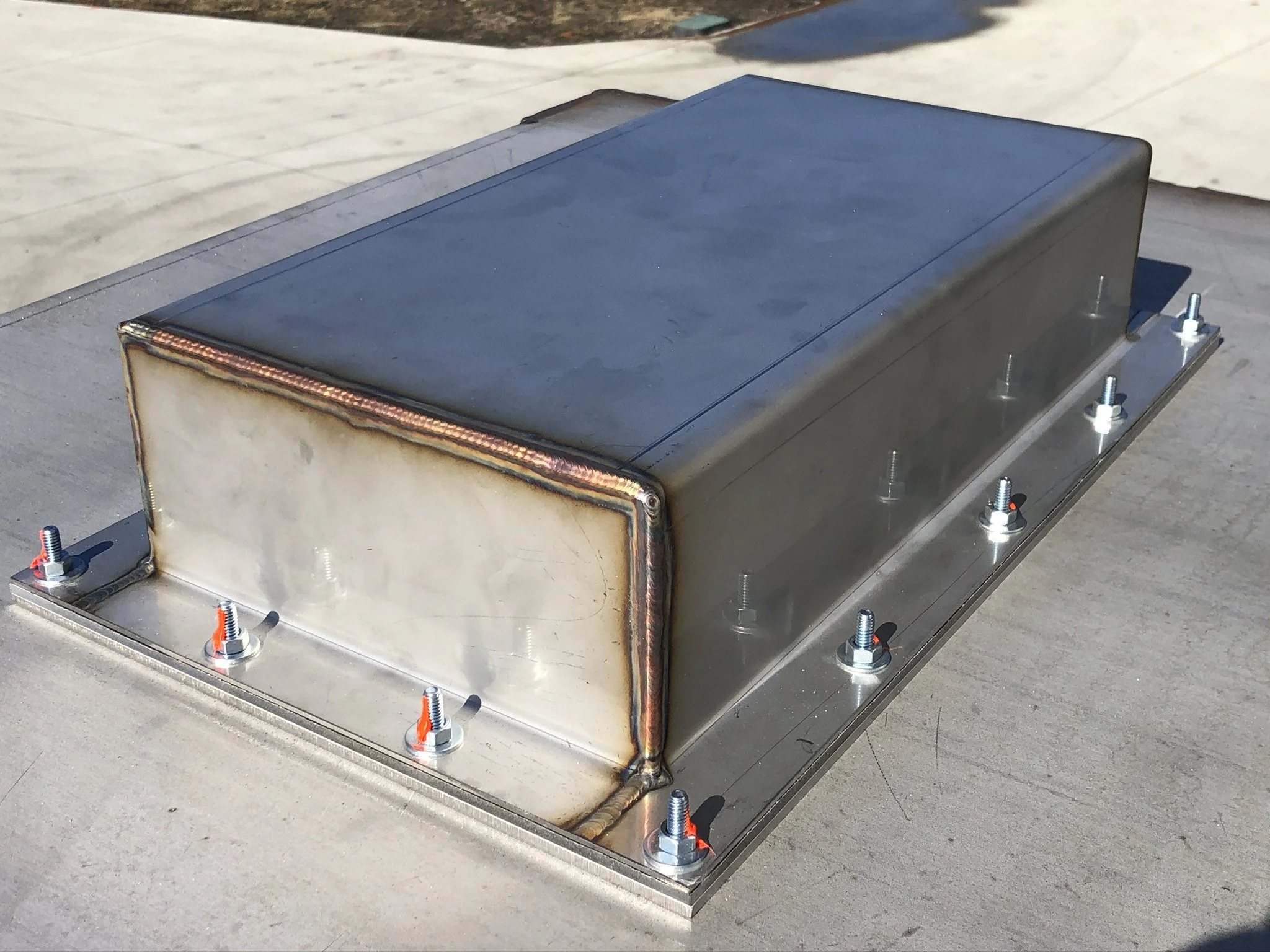 A rectangular metal electronic or mechanical enclosure mounted on a flat surface, with visible screws and bolts, featuring a weld seam along one corner.
