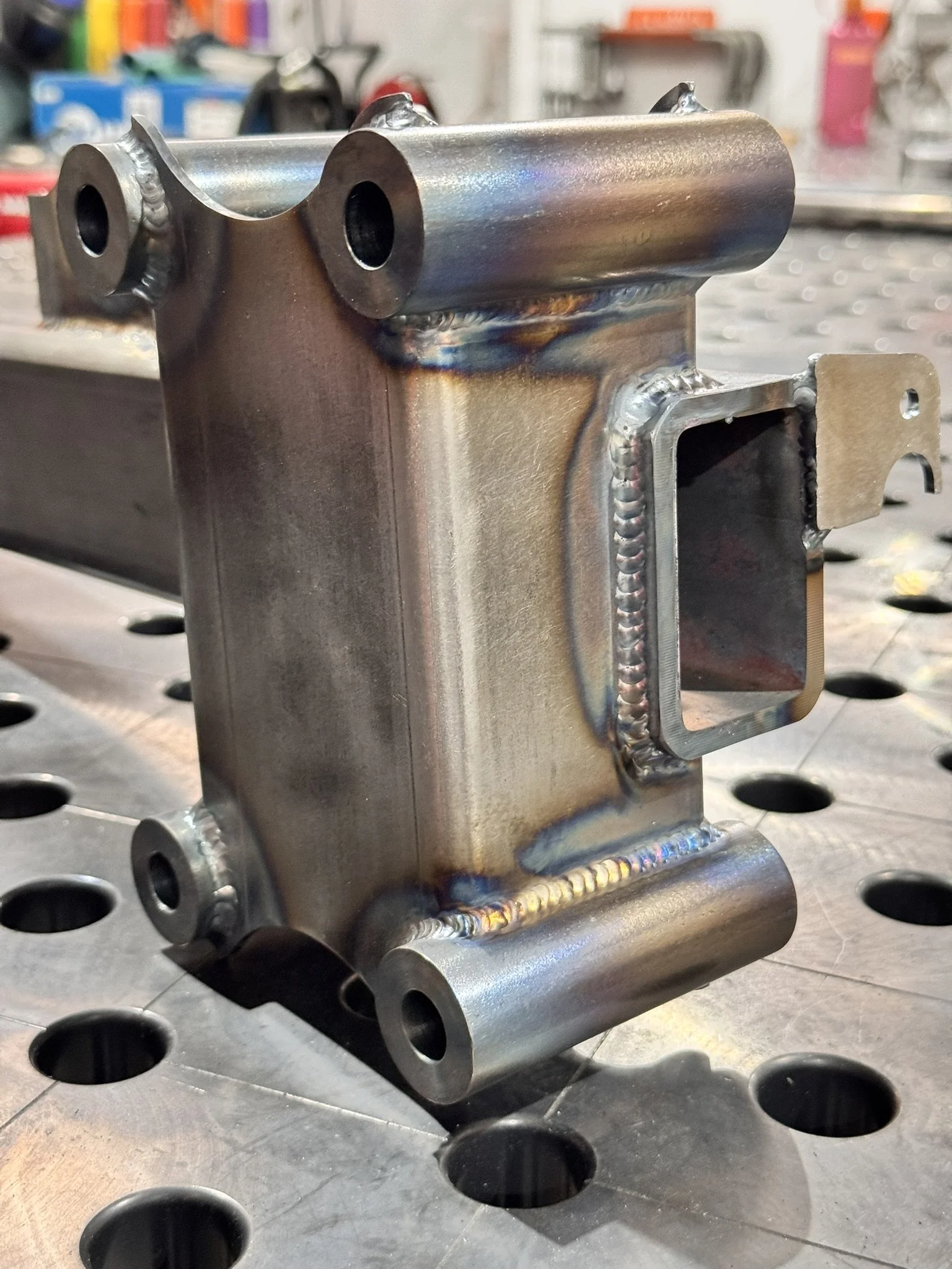 A metal welded component with a rectangular opening on one side, placed on a perforated metal work surface in a workshop setting.