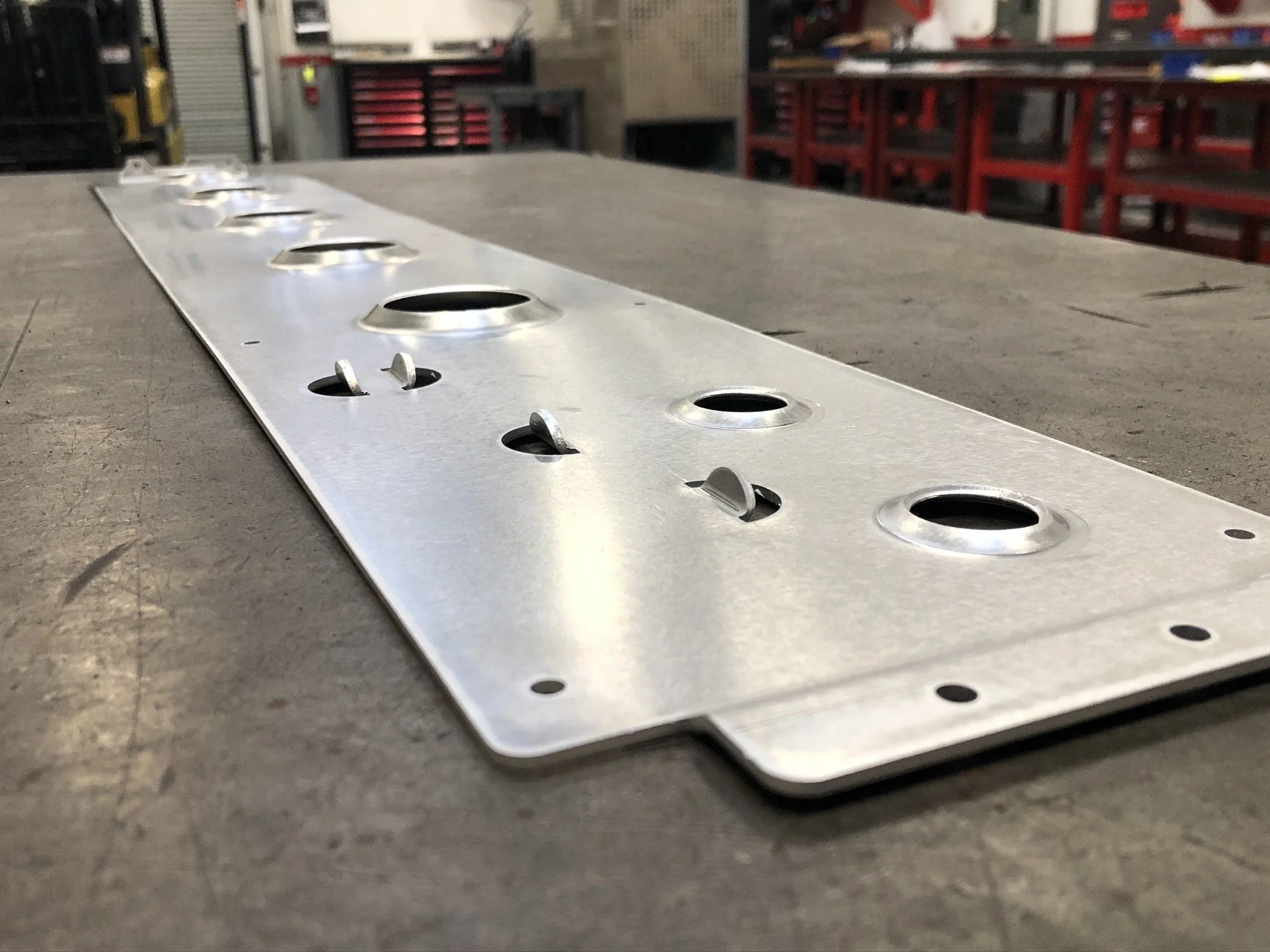 Close-up view of a metal plate with multiple round holes and small bent metal tabs, placed on a wooden work surface in an industrial workshop with red tool carts in the background.
