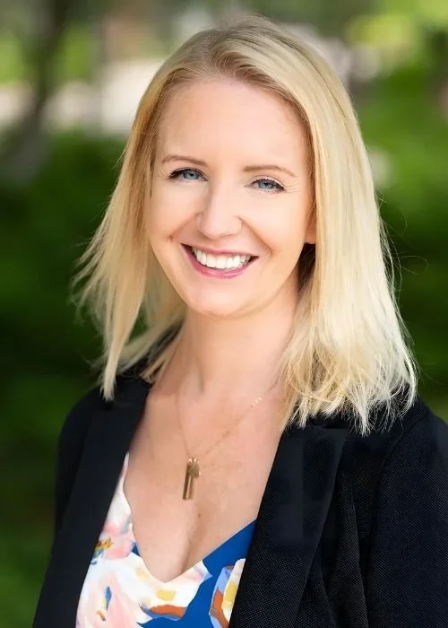 Portrait of a smiling blonde woman outdoors with green trees in the background.
