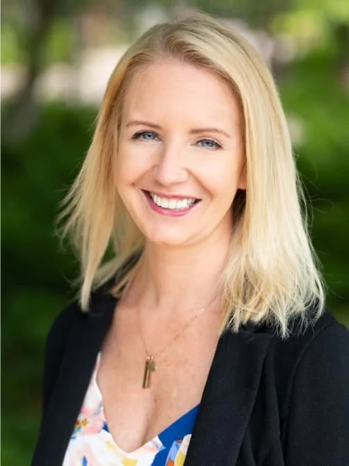 Smiling woman with blonde hair, wearing a black blazer and a colorful top, outdoors with blurred greenery in the background.