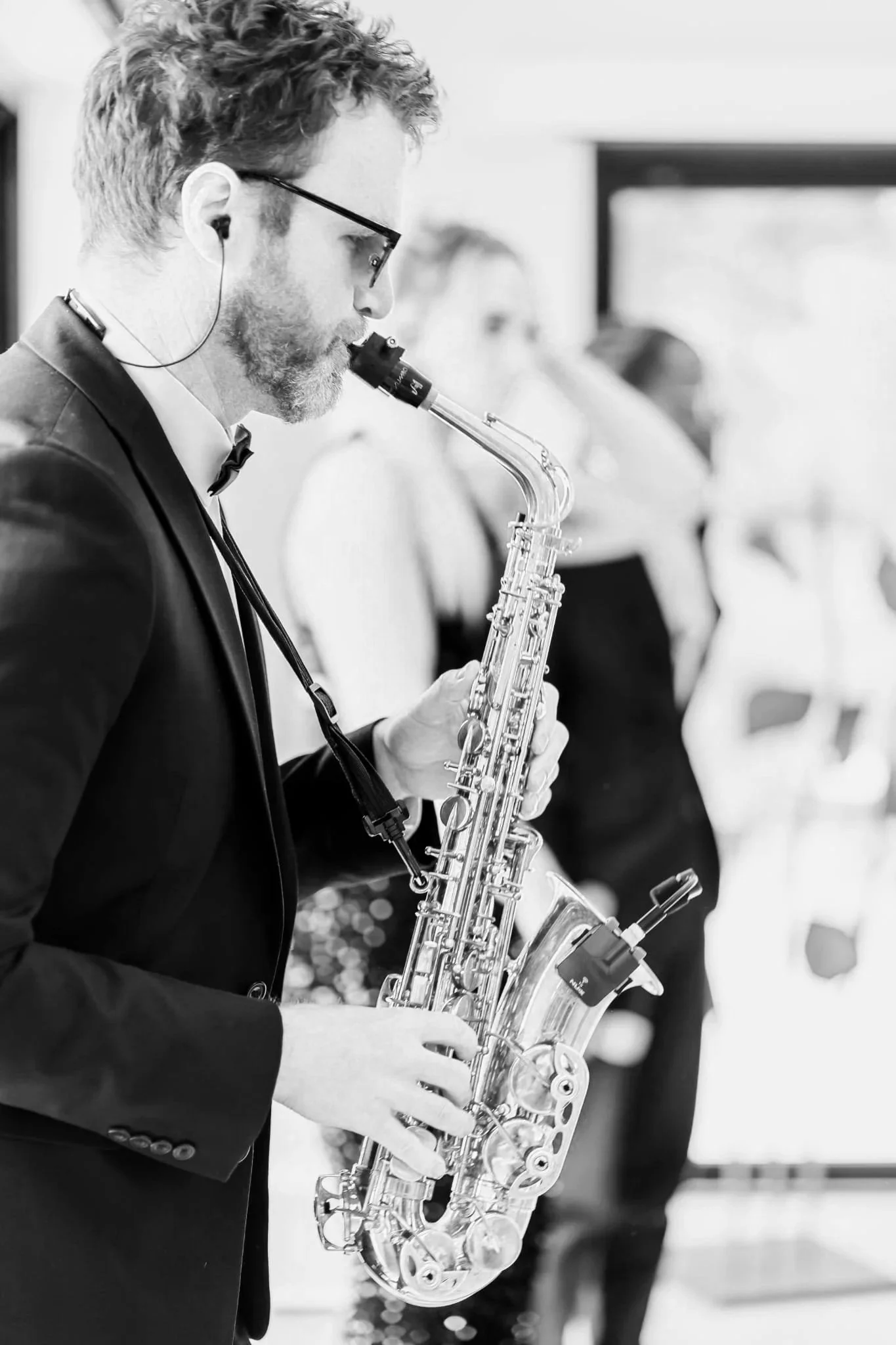 A man in a suit plays a saxophone indoors while two singers blur behind him, sunlight and greenery visible through large windows.
