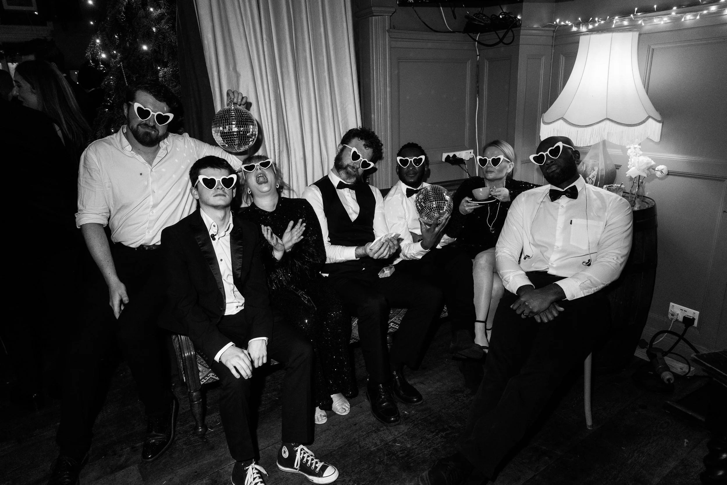 Seven adults in formalwear sit and stand together, all wearing heart-shaped sunglasses; some hold disco balls in a playful pose.