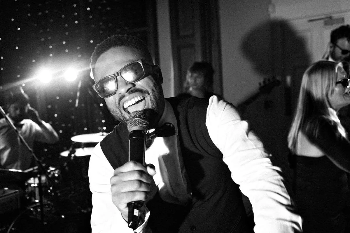Black-and-white photo of a smiling singer in sunglasses performing into a microphone, with a drummer and band members behind him in a lively venue.