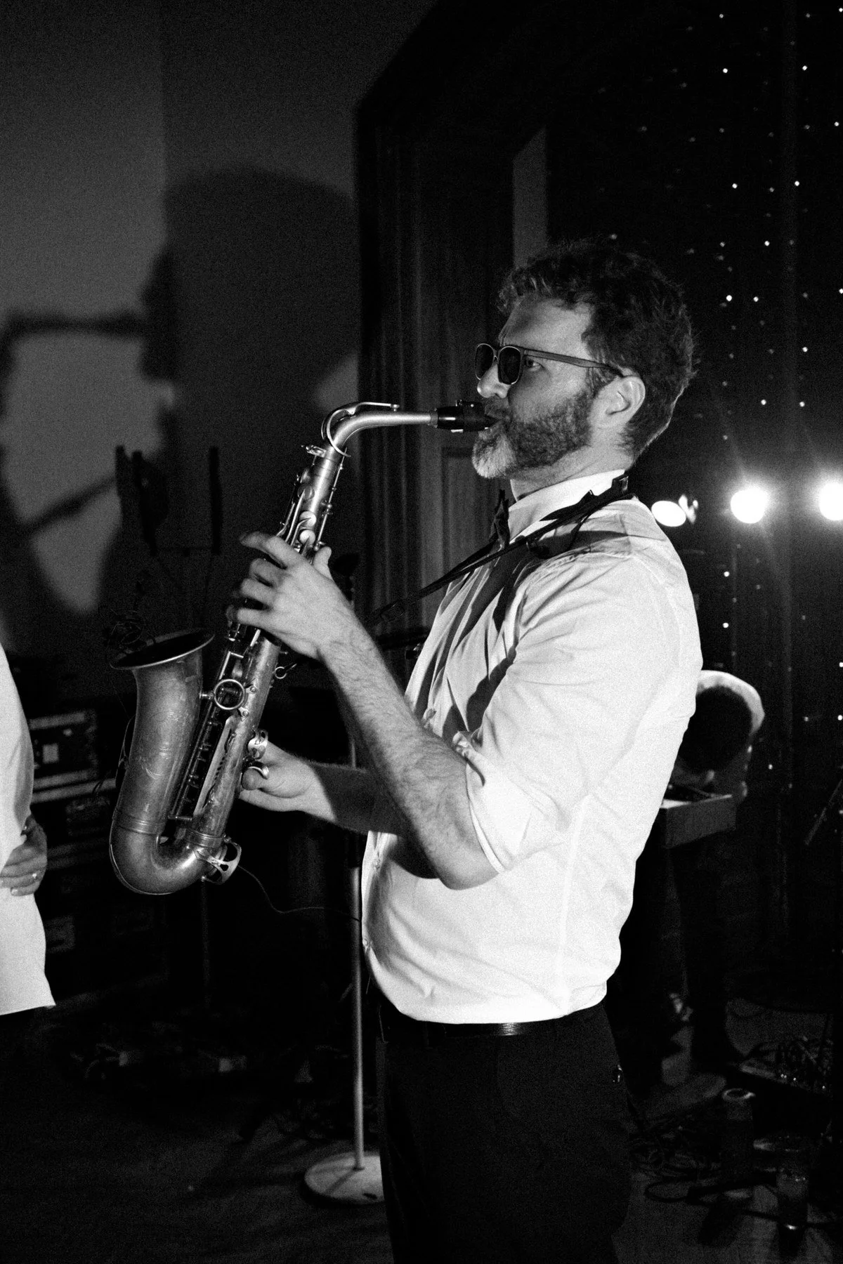 Black-and-white photo of a bearded musician in sunglasses playing saxophone onstage, surrounded by shadows, lights, and a lively concert atmosphere.