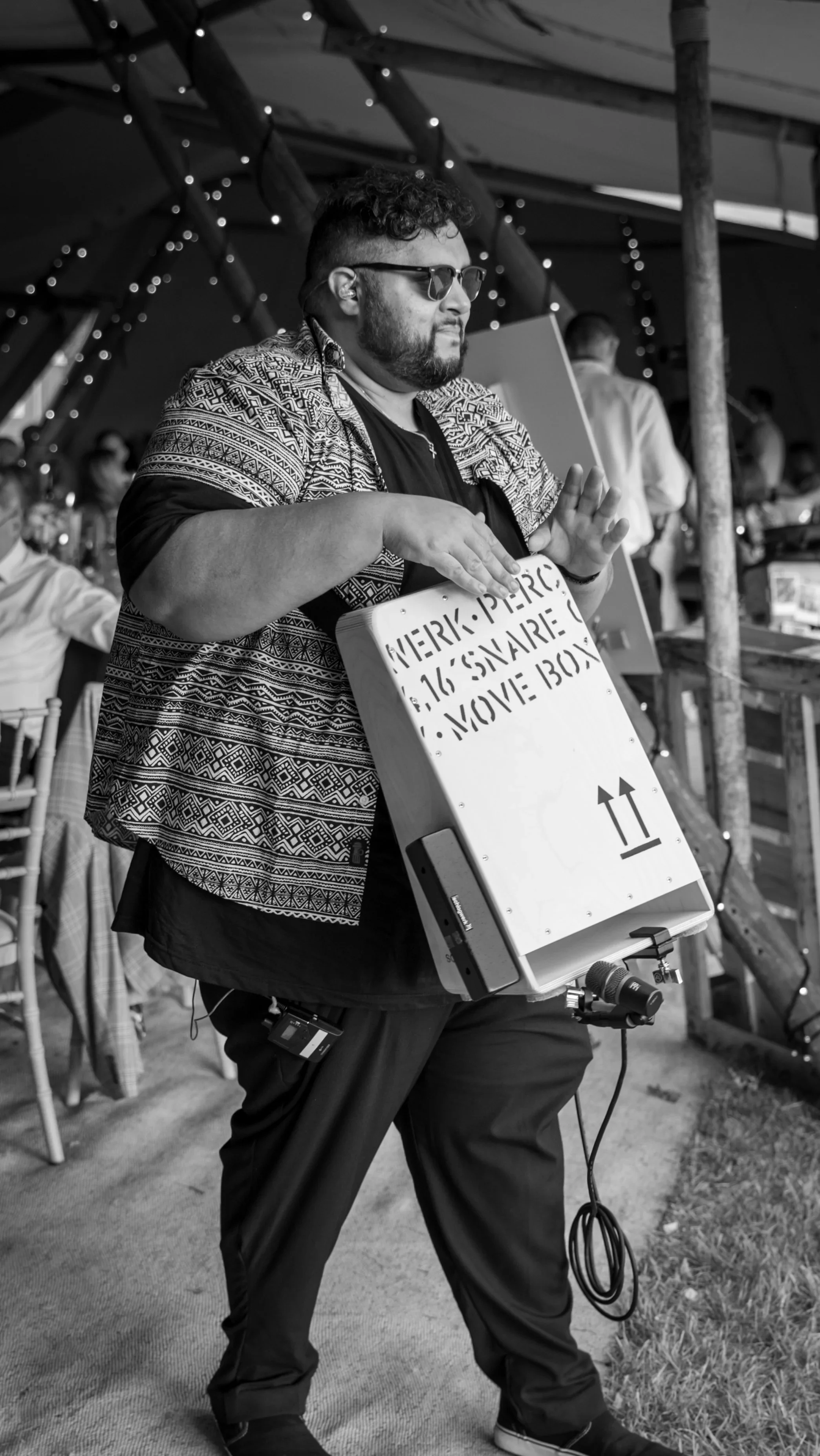A man in sunglasses plays a wearable box instrument at a wedding, tapping it like drums while guests sit under twinkling string lights in a tent.