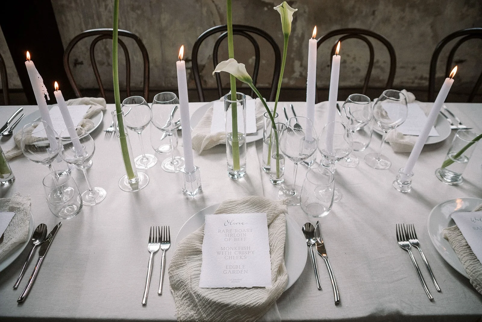 An elegant table set with white linens, glowing candles, clear glasses, and tall calla lilies, ready for a formal dinner.