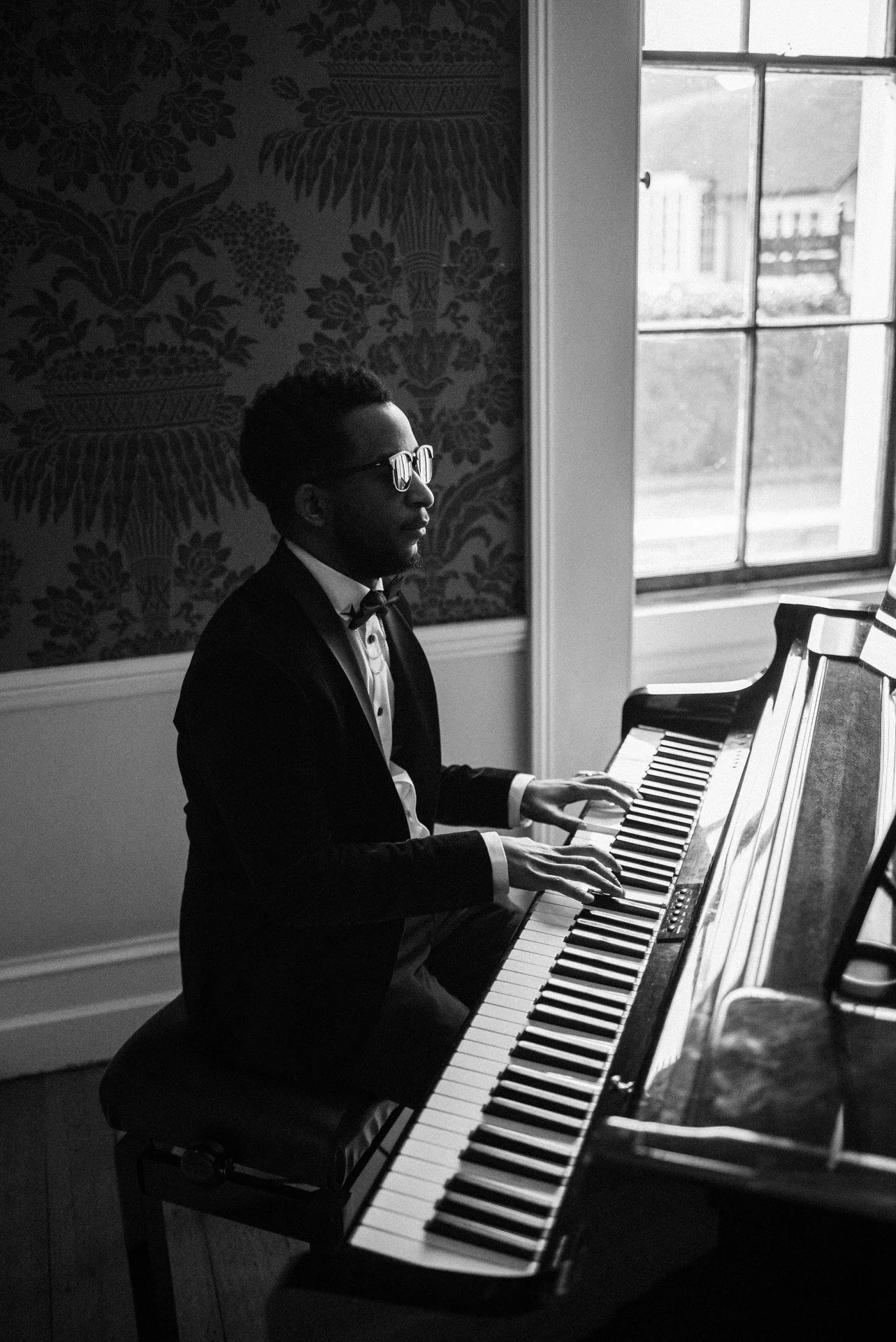A man in a tuxedo plays a grand piano by a window, light streaming in, patterned wallpaper behind him, creating a calm, elegant mood.