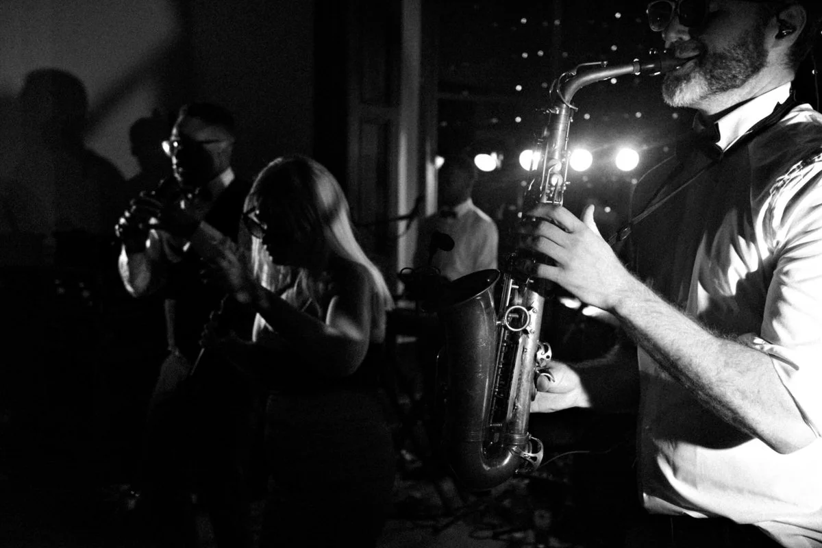 A black and white shot of a jazz band performing on stage, featuring a saxophonist in the foreground and other musicians playing in the background.