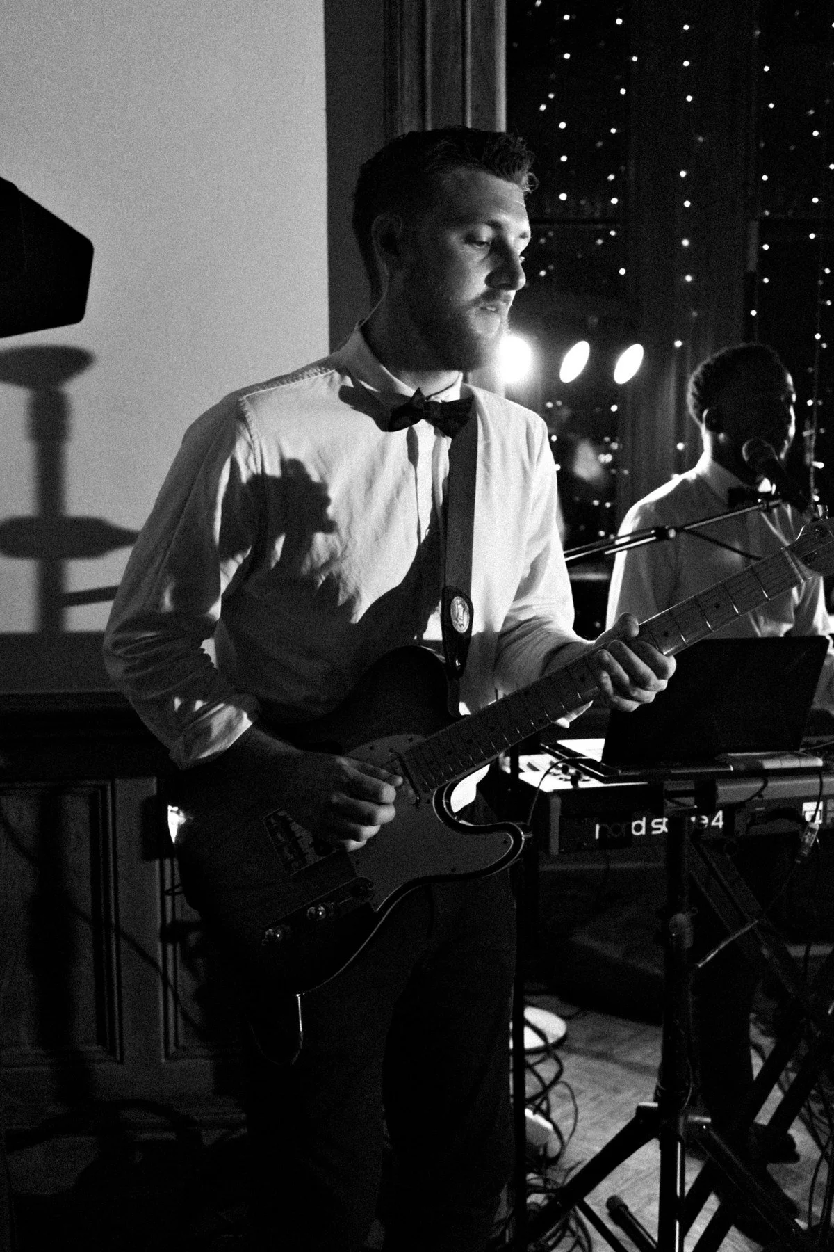 Black-and-white photo of a focused guitarist performing onstage, with a keyboardist, spotlights, and starry curtain creating a moody atmosphere.