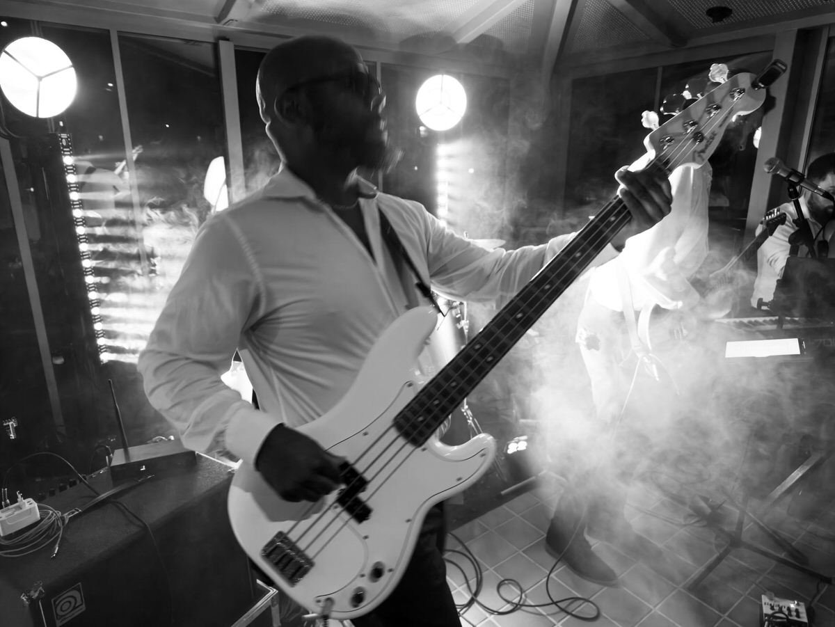 Black-and-white photo of a bassist playing a white electric bass on a smoky stage, with bright lights and another musician behind him; dramatic, energetic mood.