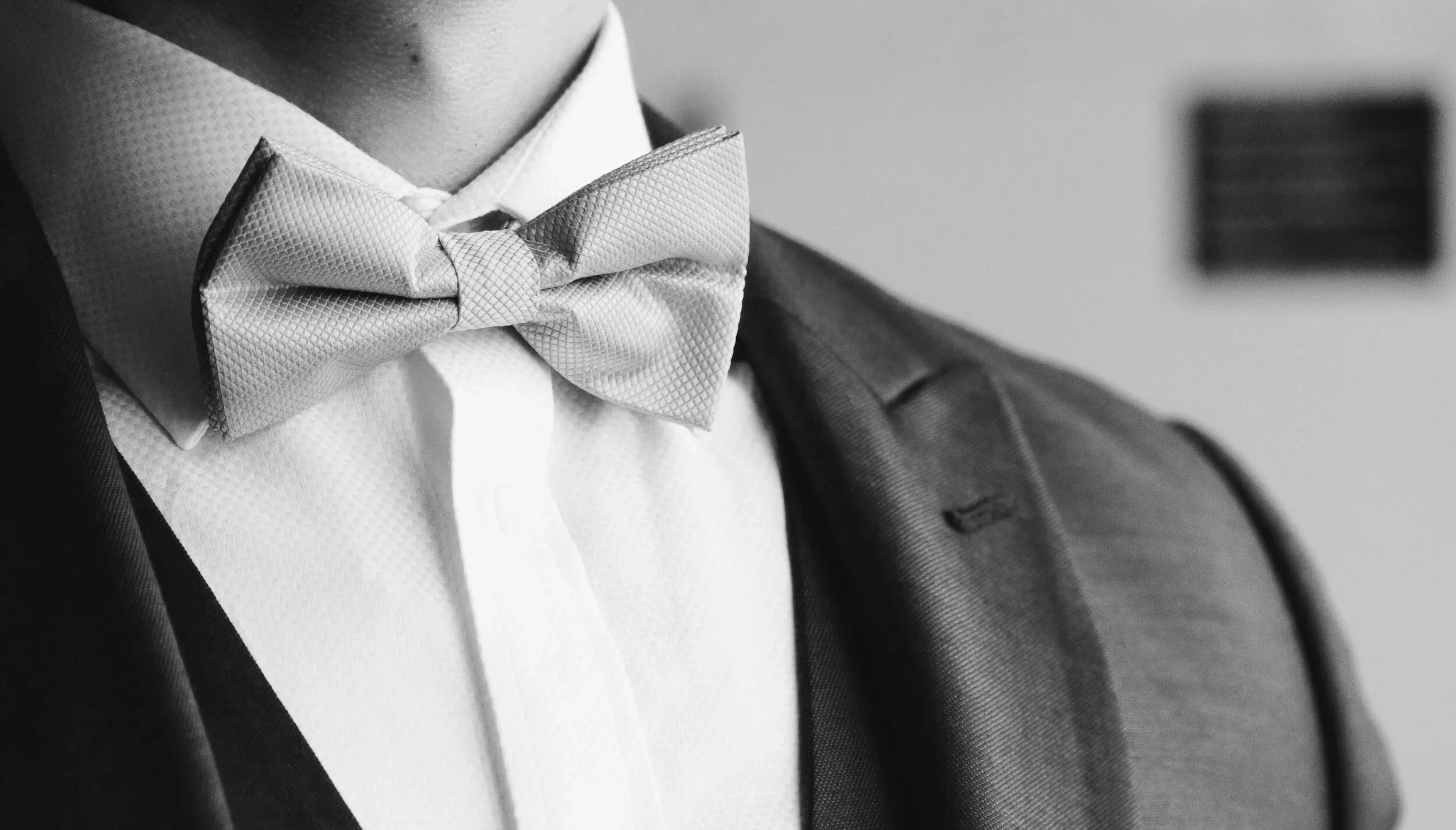 Close-up of a groom’s neck and chest: crisp white shirt, textured bow tie, and dark suit jacket catching soft light.
