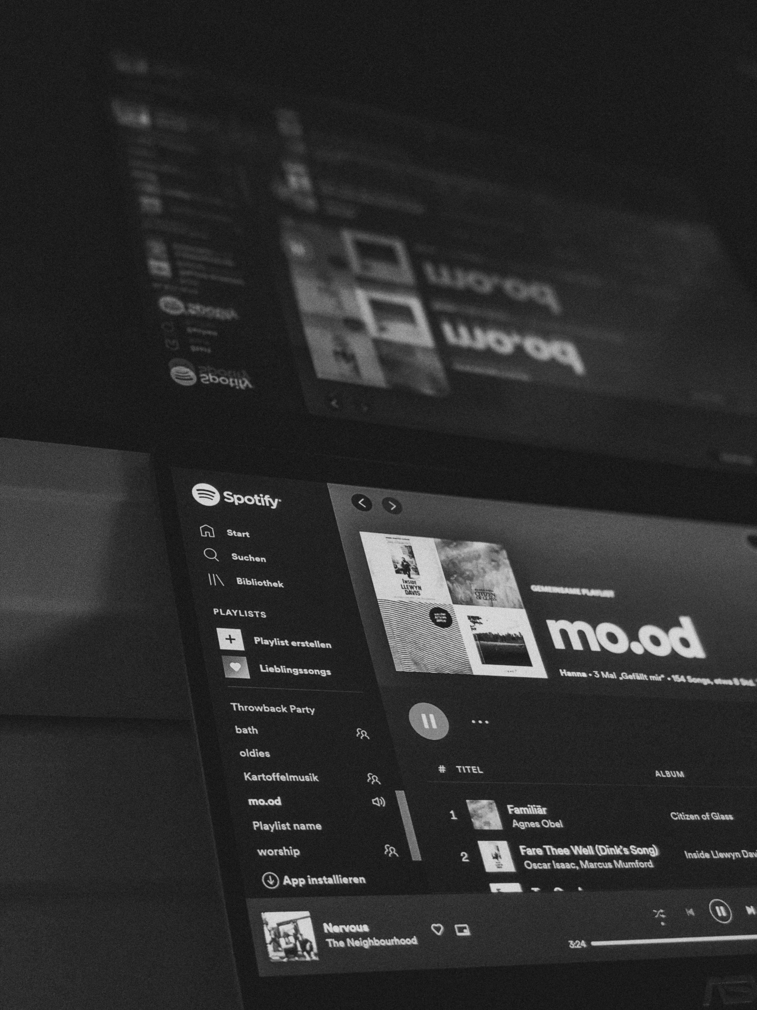 A tilted computer screen shows a music app playlist and song list in grayscale, with reflections across the glass and soft shadows around the edges.