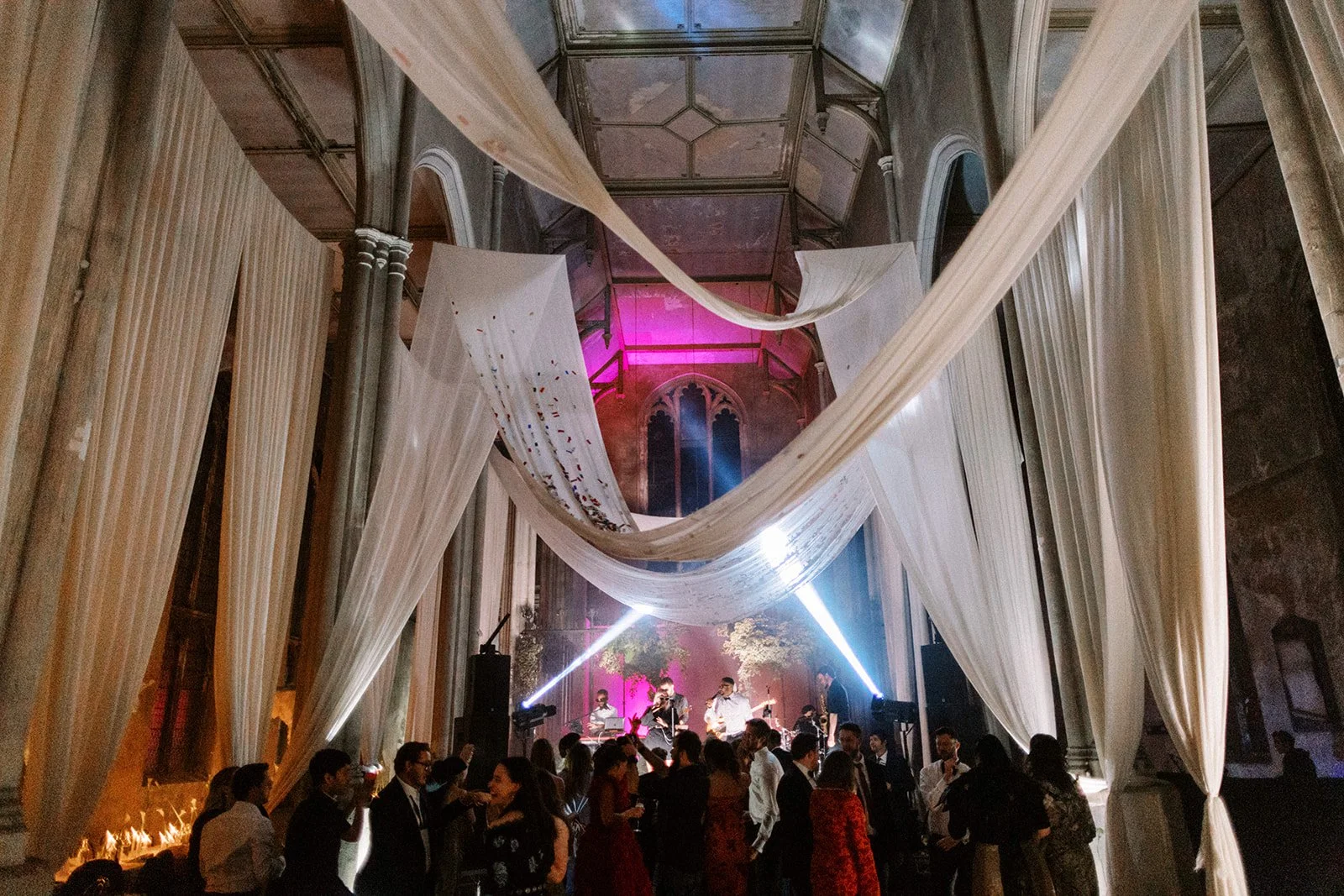 Guests dance beneath sweeping white drapes in a grand hall, as a live band plays under pink and white stage lights at the far end.
