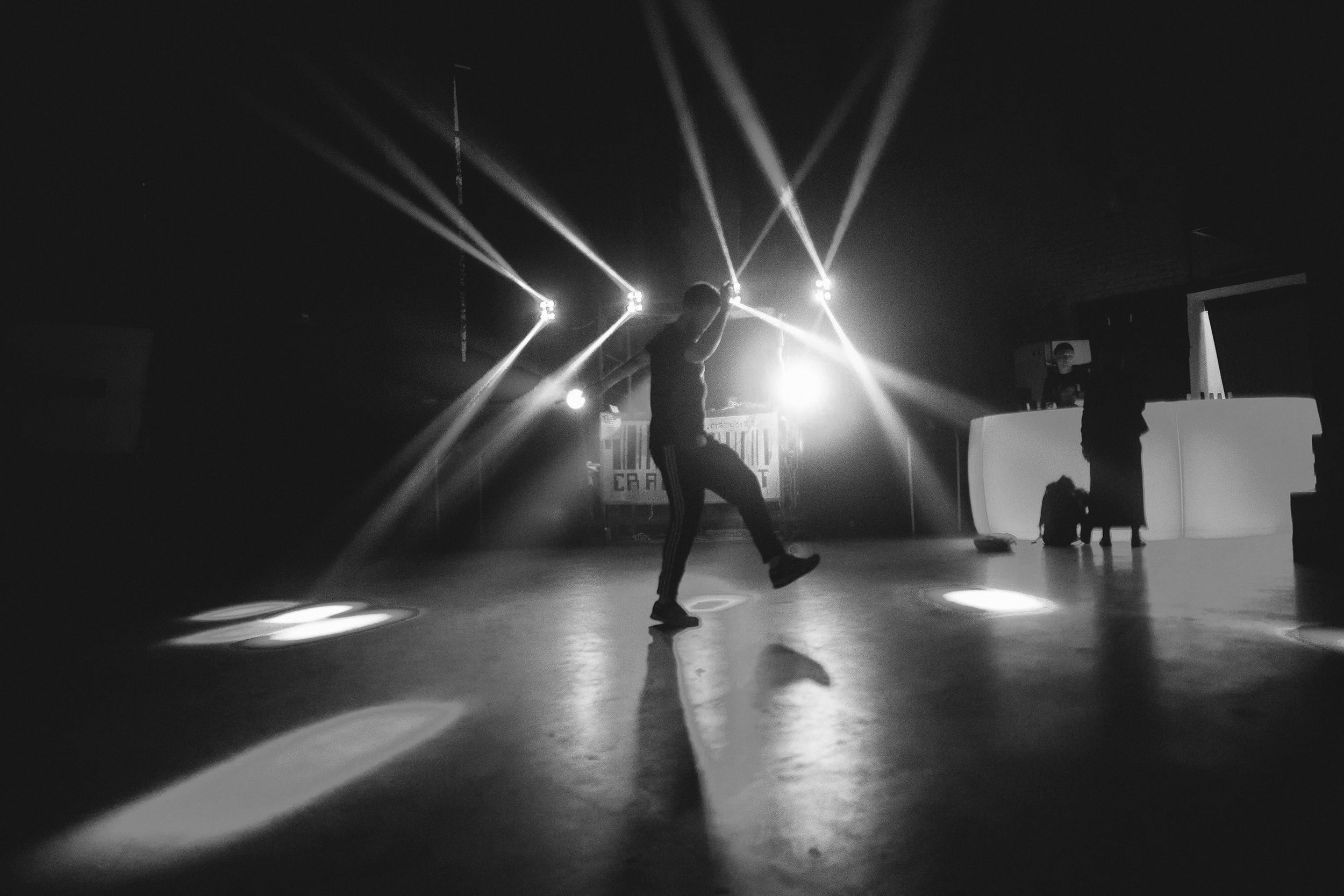 A lone dancer appears in silhouette on a glossy floor, spotlights slicing the darkness behind them and casting sharp reflections across the stage.