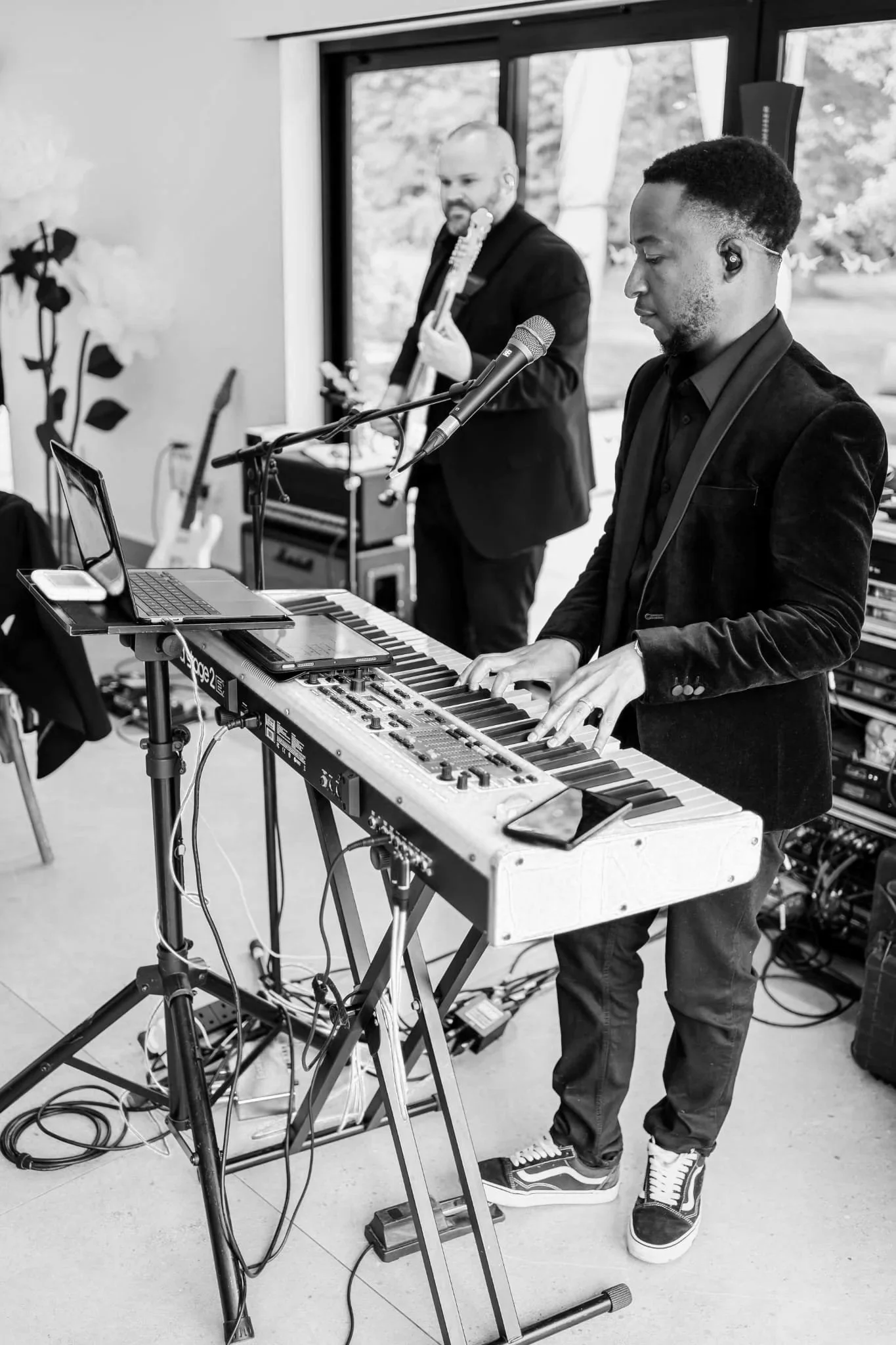 A suited keyboardist plays a red electric piano indoors, with cables and gear around him while a guitarist stands behind near bright windows.