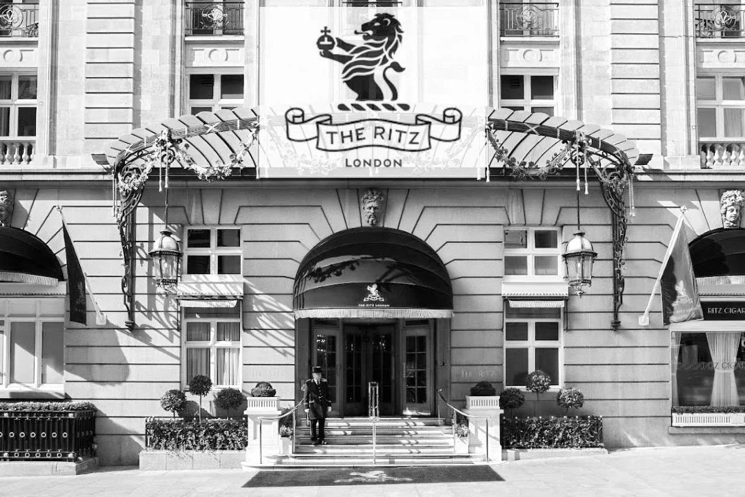 Black-and-white view of The Ritz London entrance, with ornate iron canopy, the Ritz crest above the doors, flags, and a uniformed doorman standing outside.