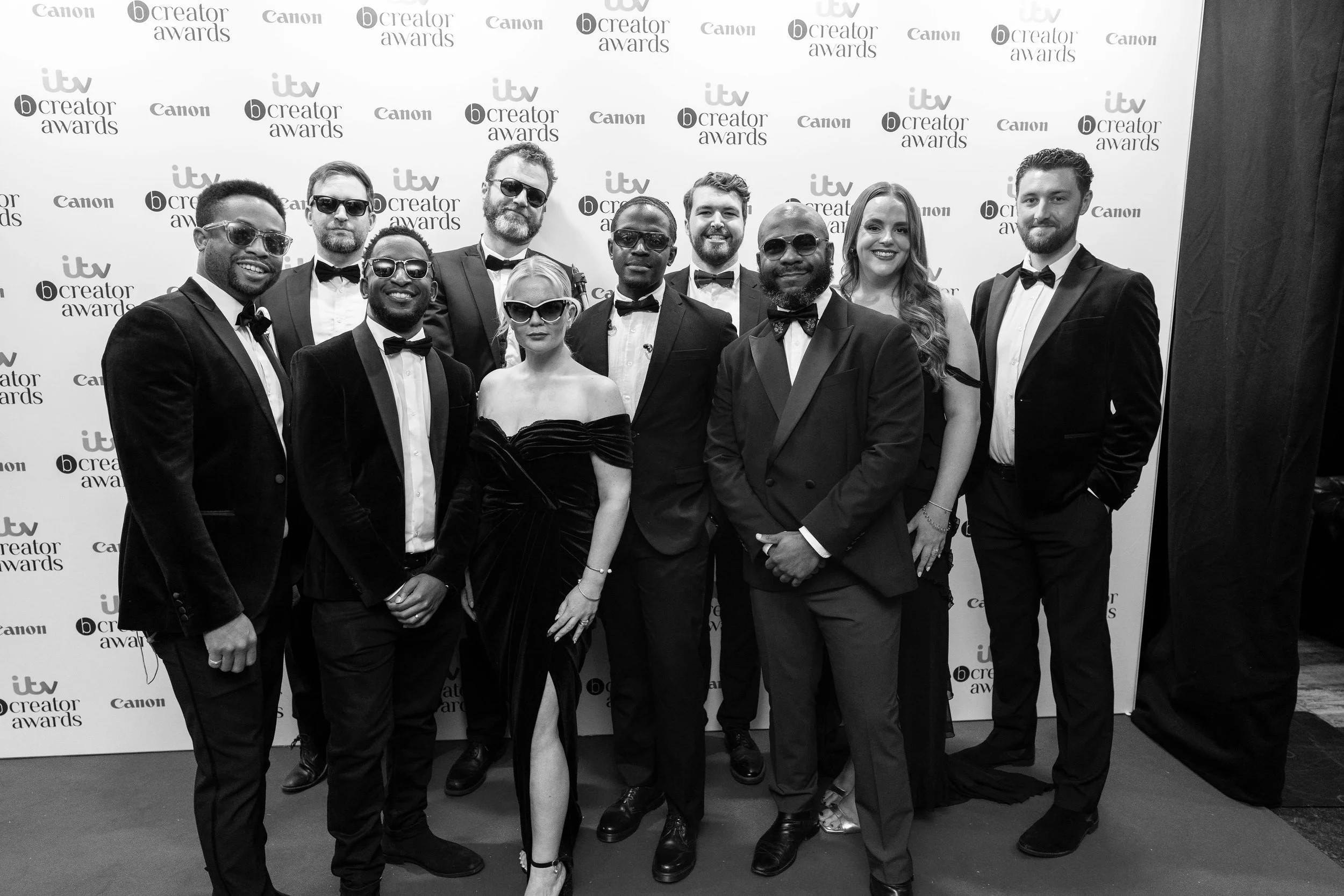 A group in tuxedos and gowns pose on a red carpet before a branded backdrop, smiling and wearing sunglasses at a formal awards event.