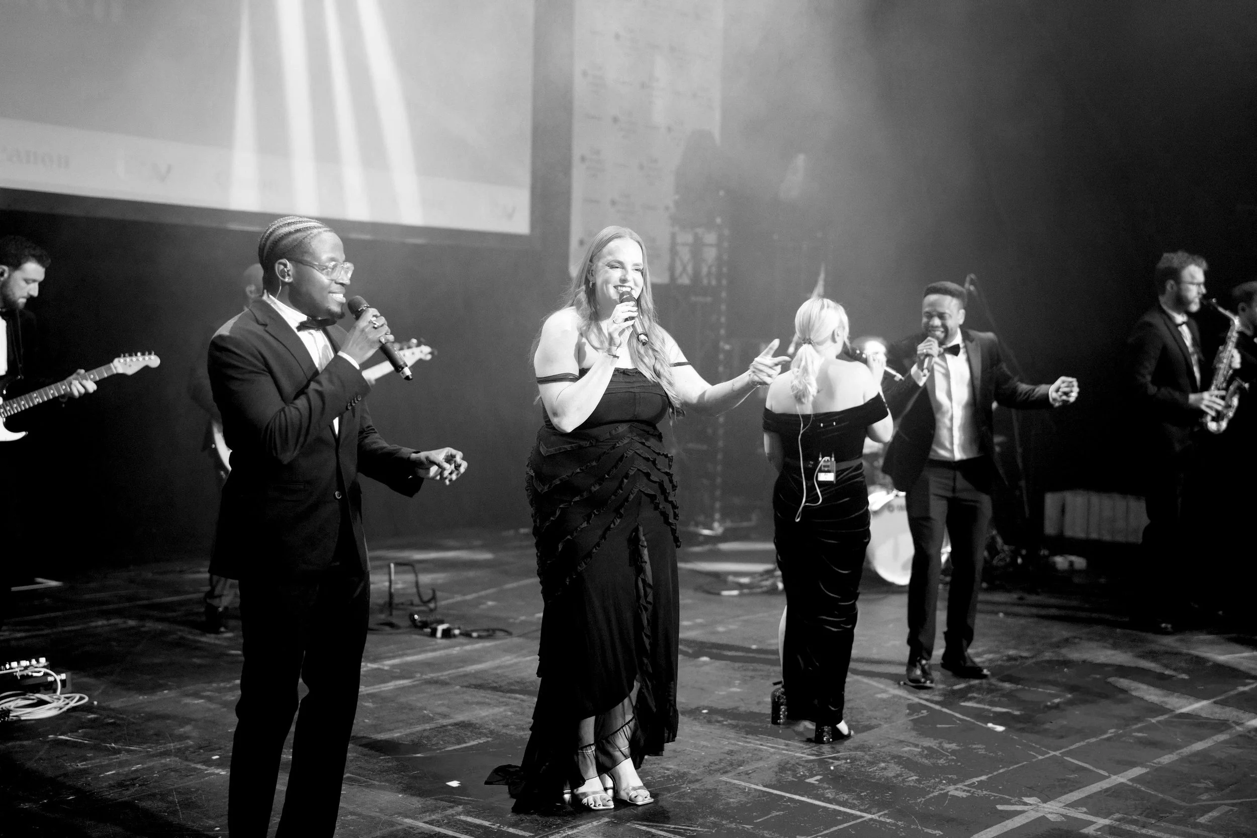 NeoRoots vocalists sing together on stage at a London awards event, performing live with the band and horn section behind them.