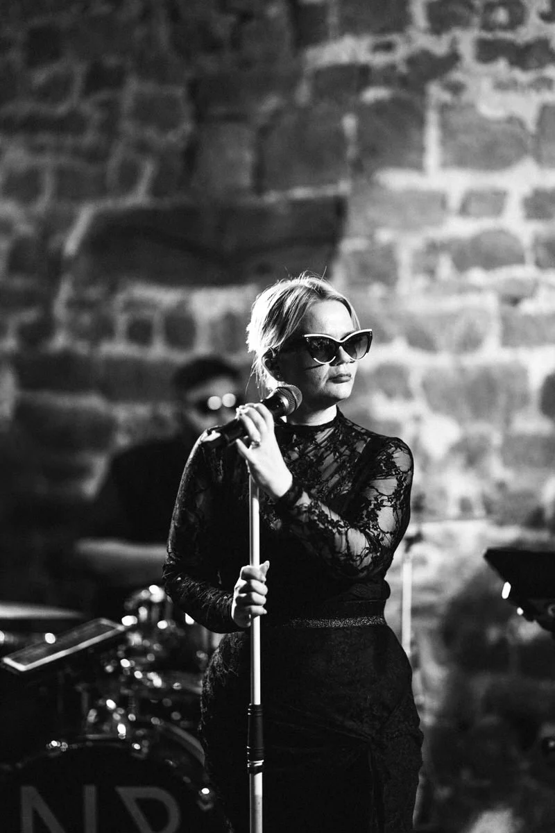 A singer in a sparkly short dress and sunglasses performs on stage with a full band behind her, lights glowing as musicians line up and play.