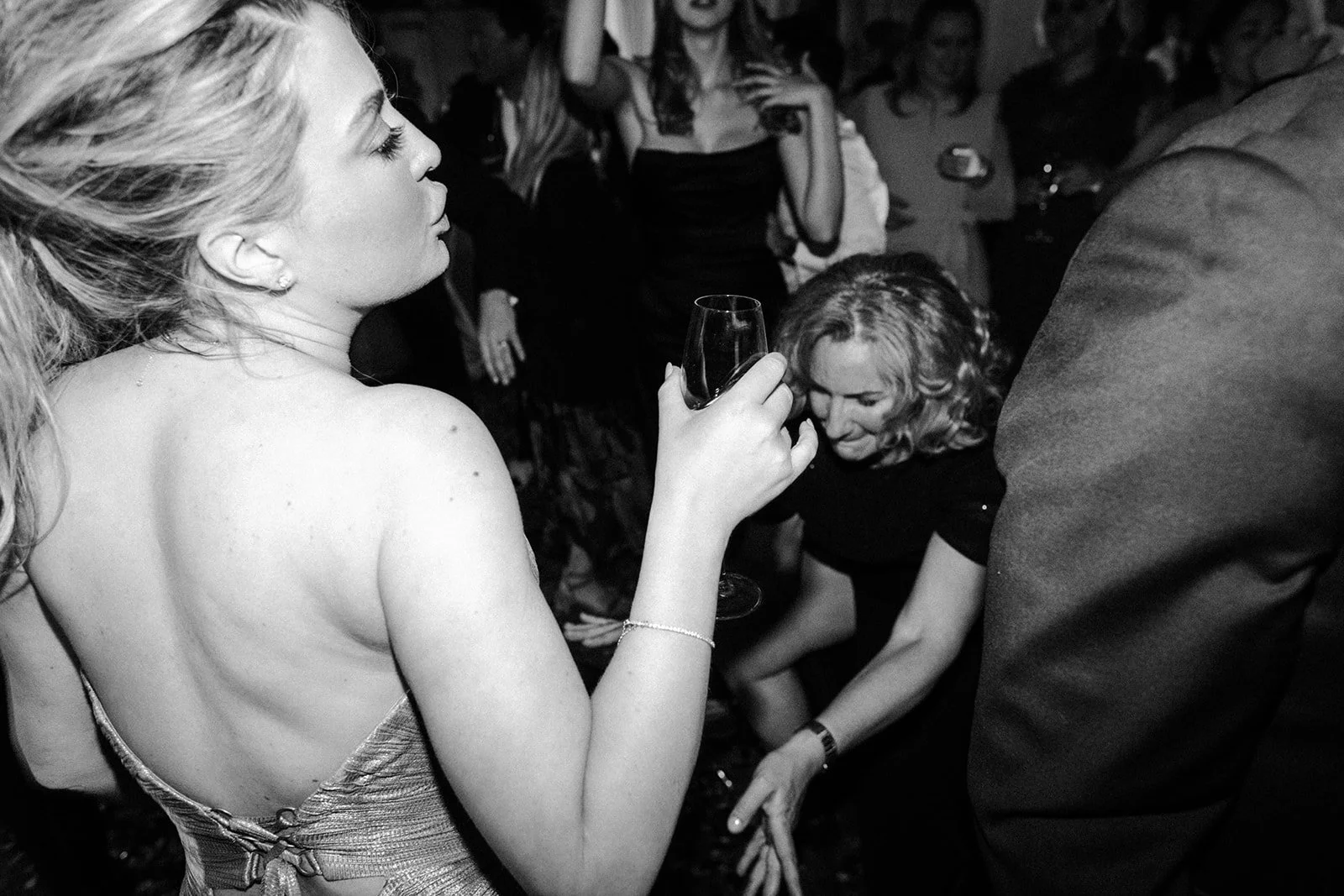 A woman in a backless dress holds a wine glass on a crowded dance floor, guests moving and celebrating around her.