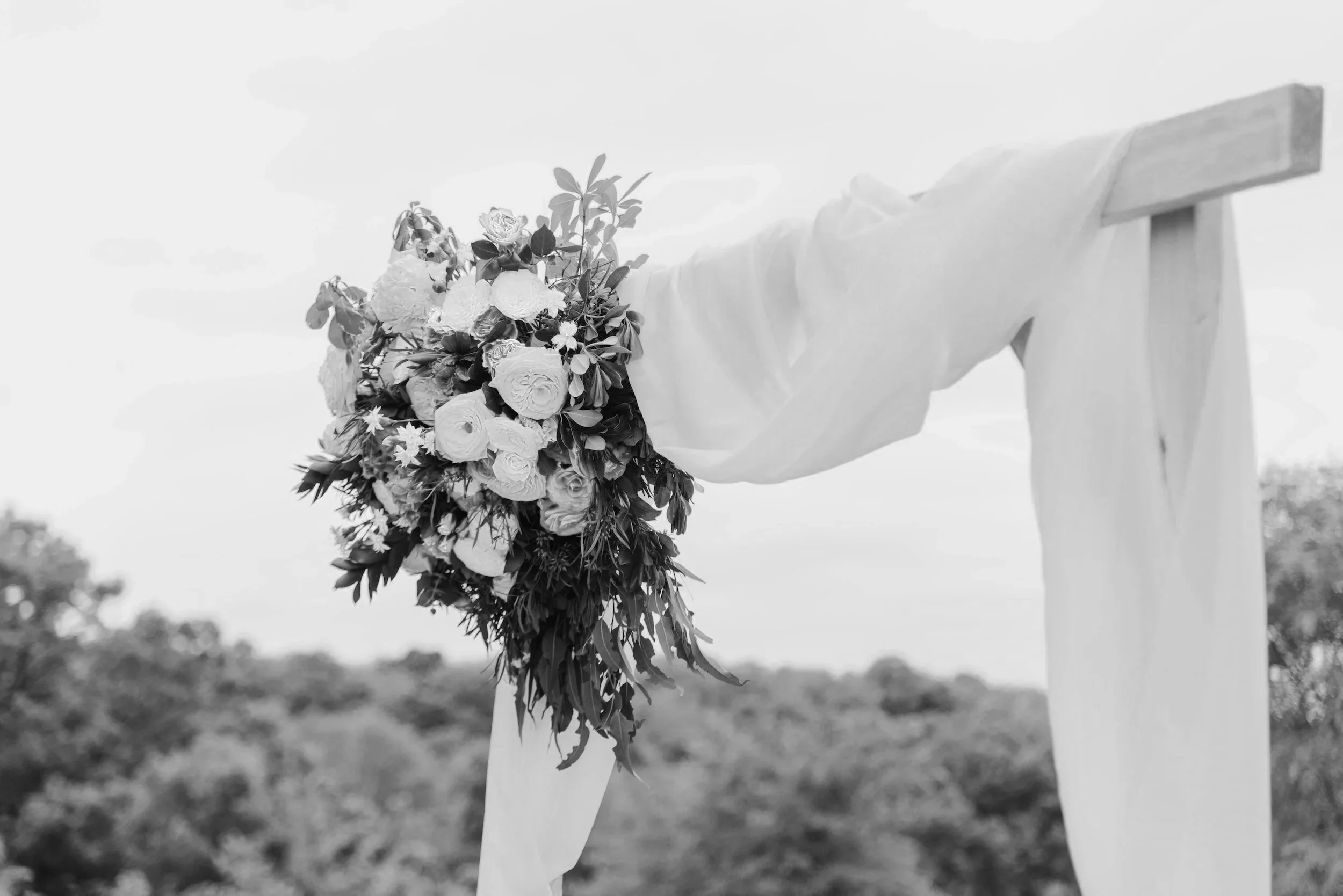 A lush bouquet hangs from a wooden arch draped in soft fabric, standing outdoors against a calm sky and distant trees, ready for a wedding ceremony.