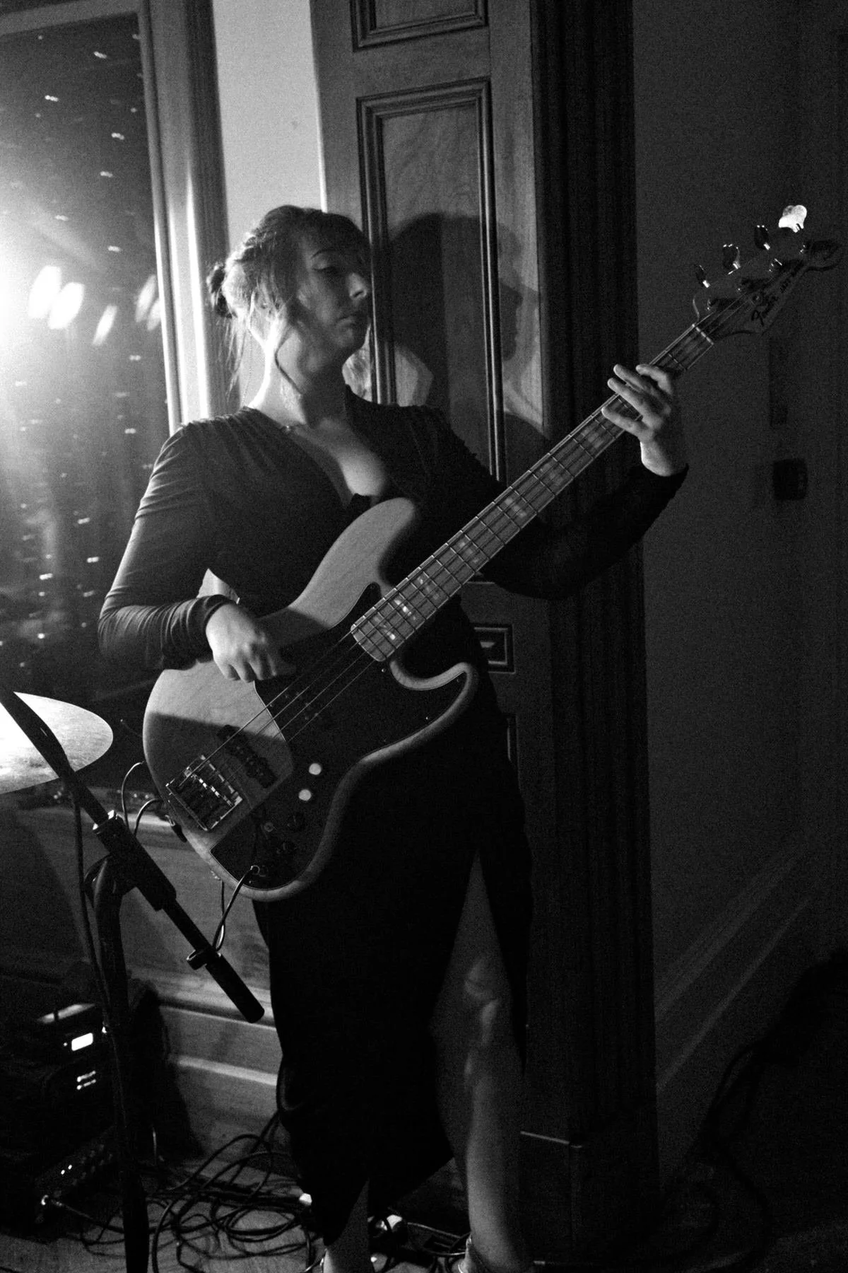 Black-and-white photo of a woman playing electric bass in a dim room, backlit by a window, with drum hardware and cables nearby, casting a dramatic mood.