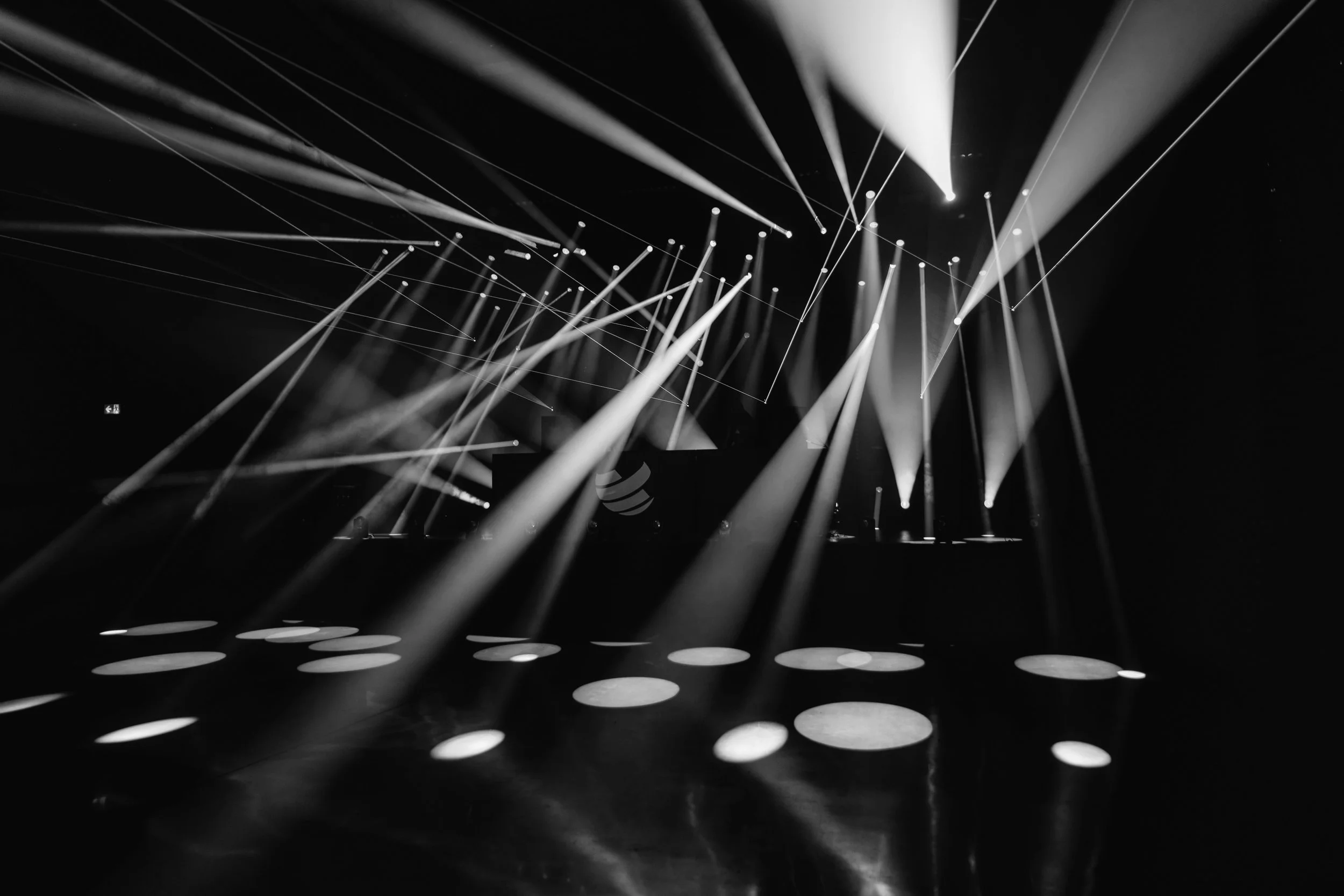A dark stage filled with sweeping white beams and sharp light rays, lasers crossing overhead while pale circles glow across the floor.