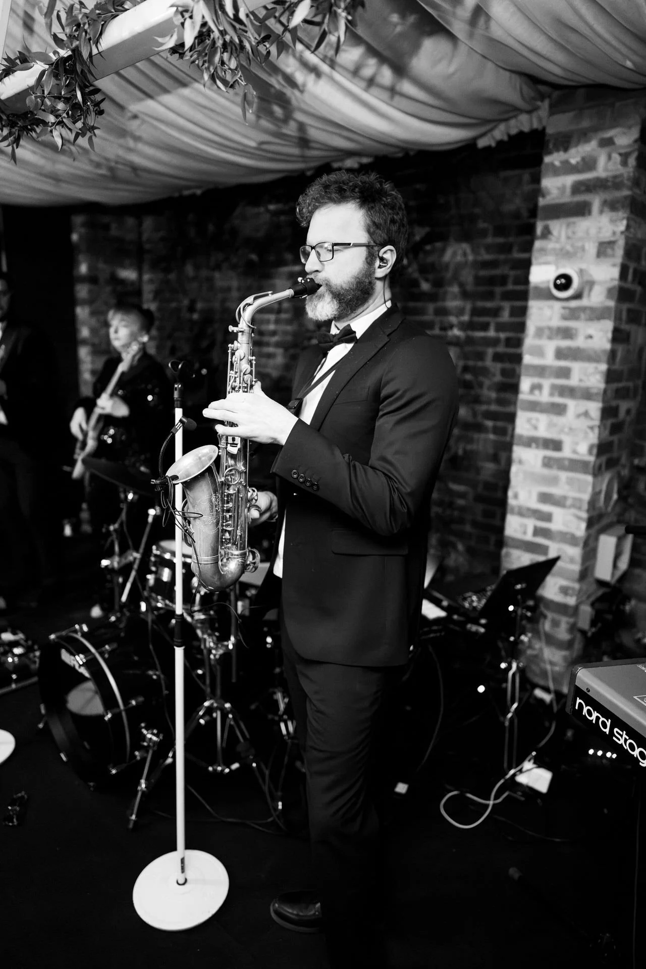 A tuxedoed saxophonist plays under draped fabric and leafy décor, brick walls behind him, with bandmates and drums fading softly into the background.