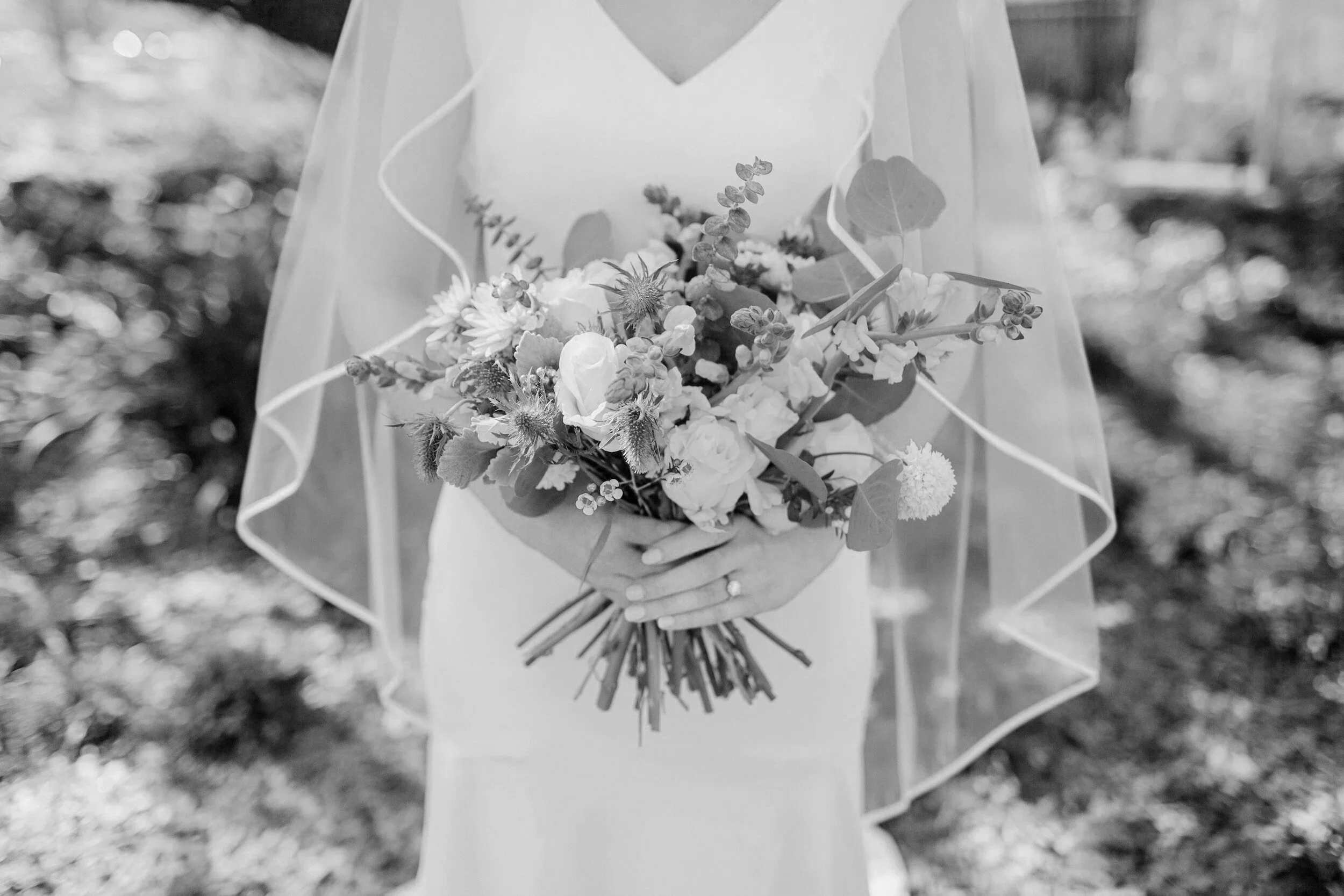 A bride in a simple gown and veil holds a full bouquet of mixed flowers at her waist, hands gently clasped, standing outdoors in soft light.