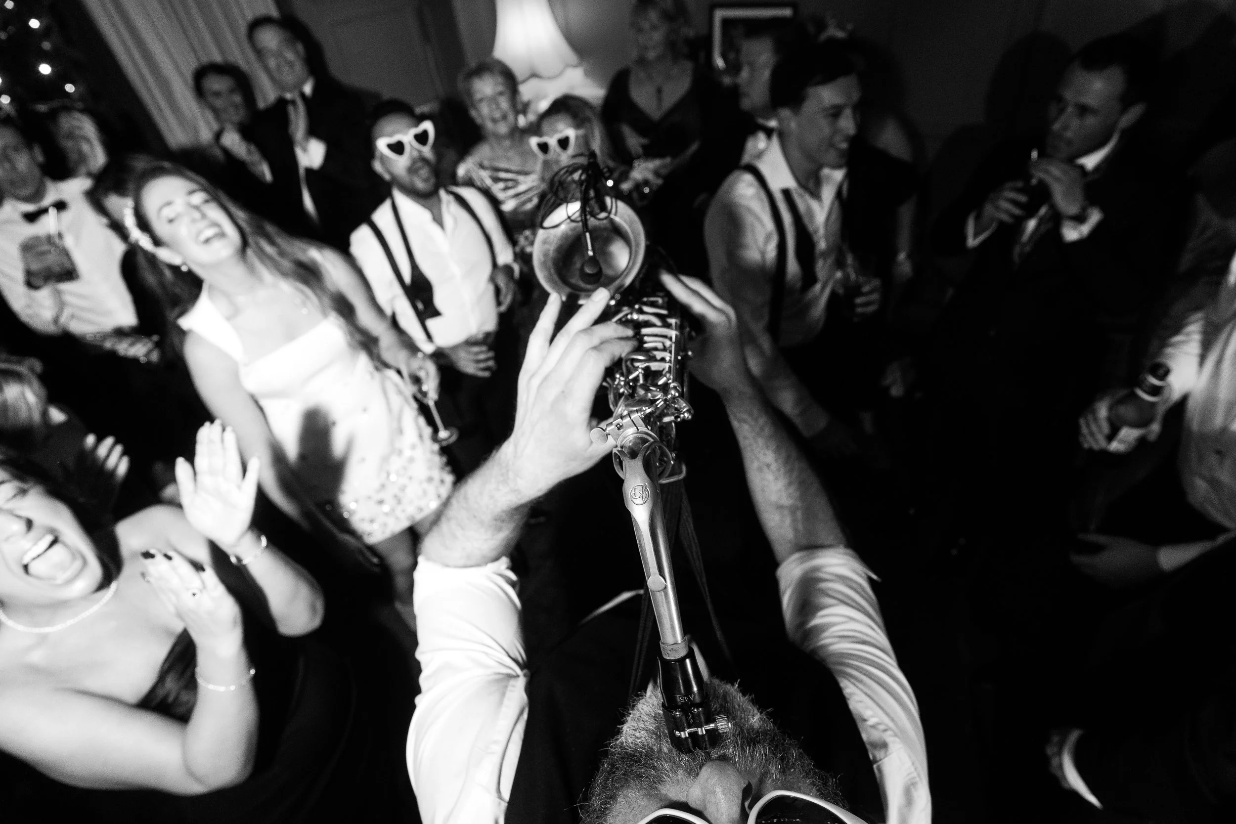 Black-and-white shot from behind a saxophonist playing into a cheering, tightly packed crowd on a lively dance floor.
