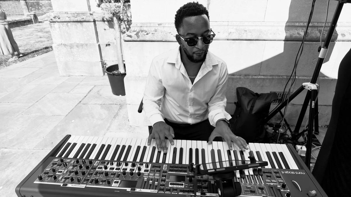 A man with a beard and sunglasses wearing a white button-down shirt plays a Nord Stage 4 keyboard outdoors on a sunny day in this black and white photo.