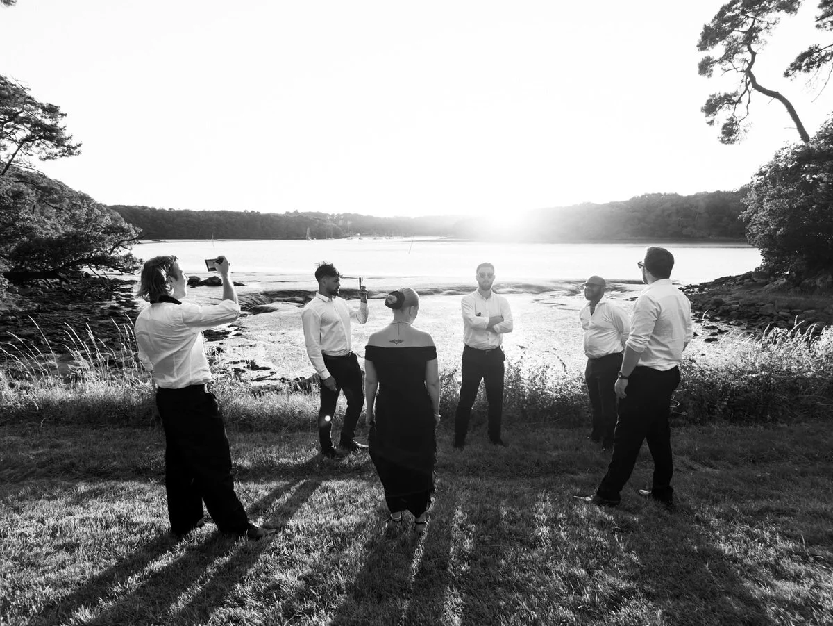 Black-and-white photo of six people posing by a lake at sunset, with one person taking a picture; long shadows and calm scenery create a reflective mood.