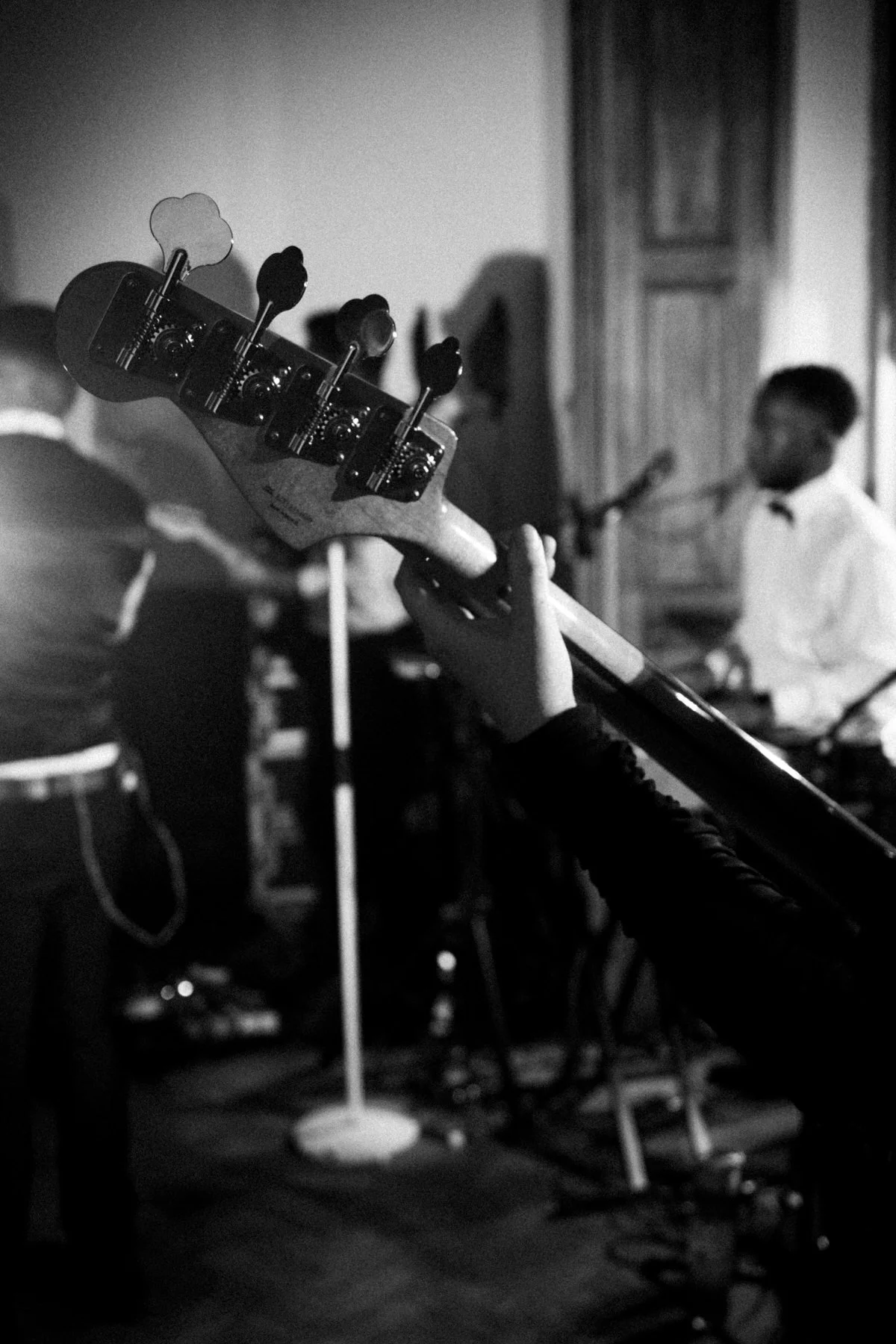 Black-and-white live music scene: a musician’s hand grips a bass guitar in the foreground while a band performs in a dim room.