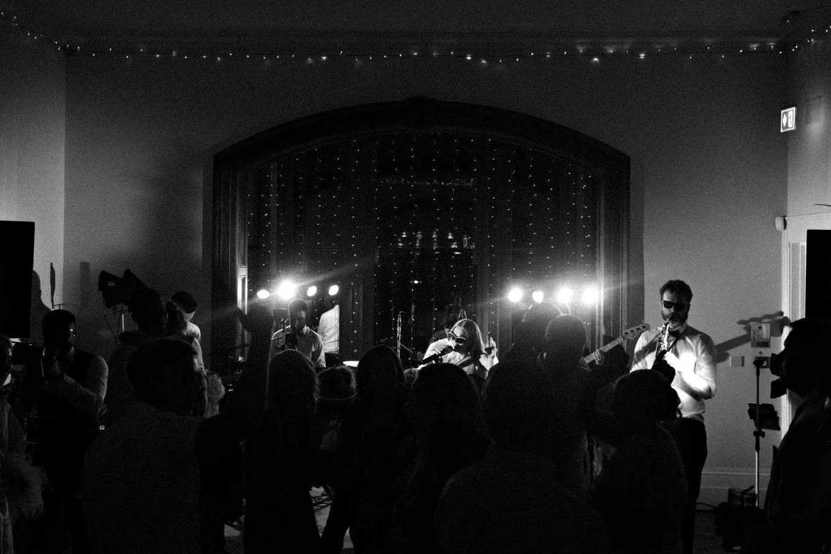 A black and white scene shows a band performing on a stage with string lights in the background, while a crowd dances in the dimly lit foreground.