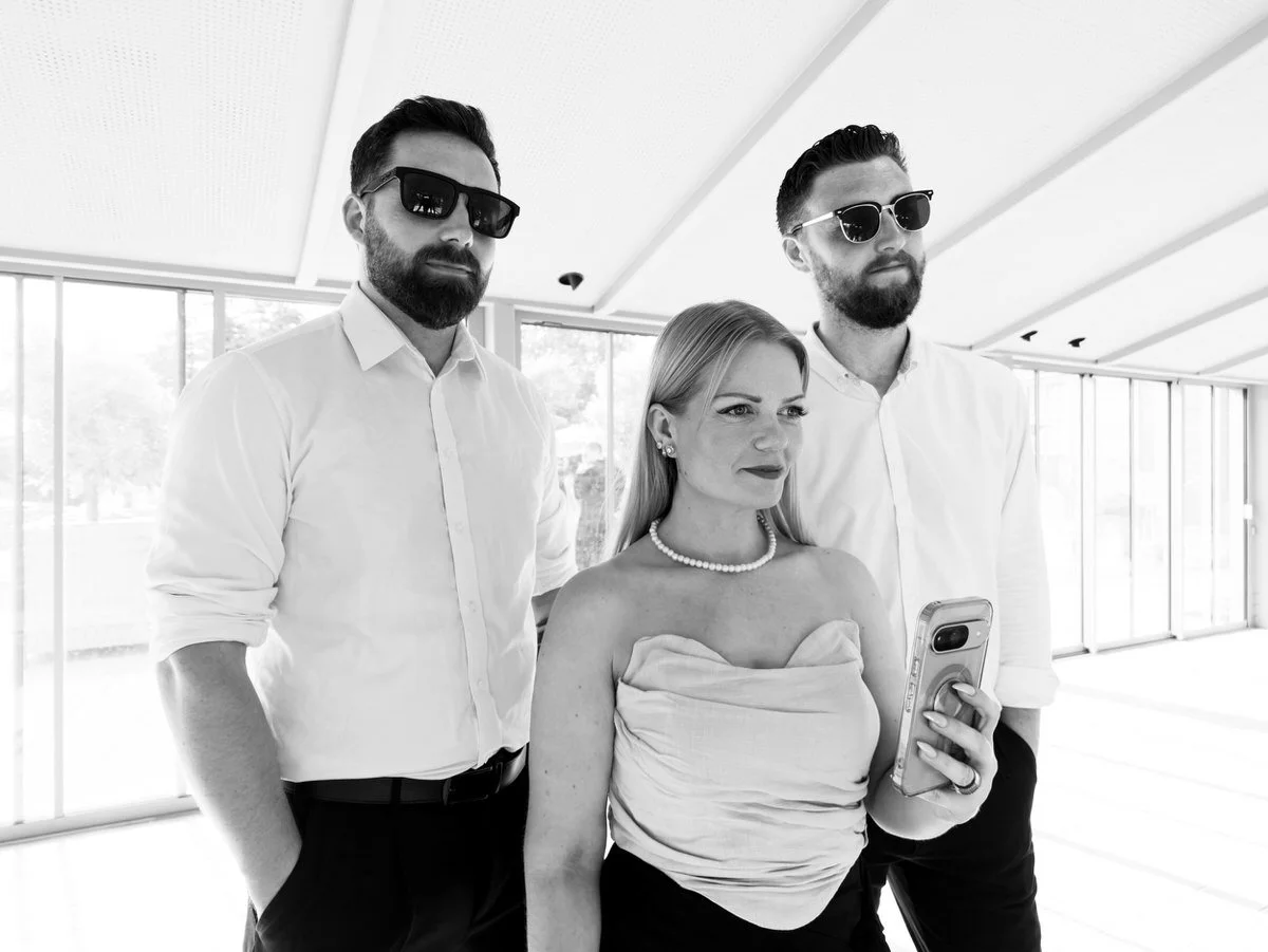 A poised woman holds her phone between two bearded men in sunglasses in a bright modern interior, creating a cool, confident monochrome fashion portrait.