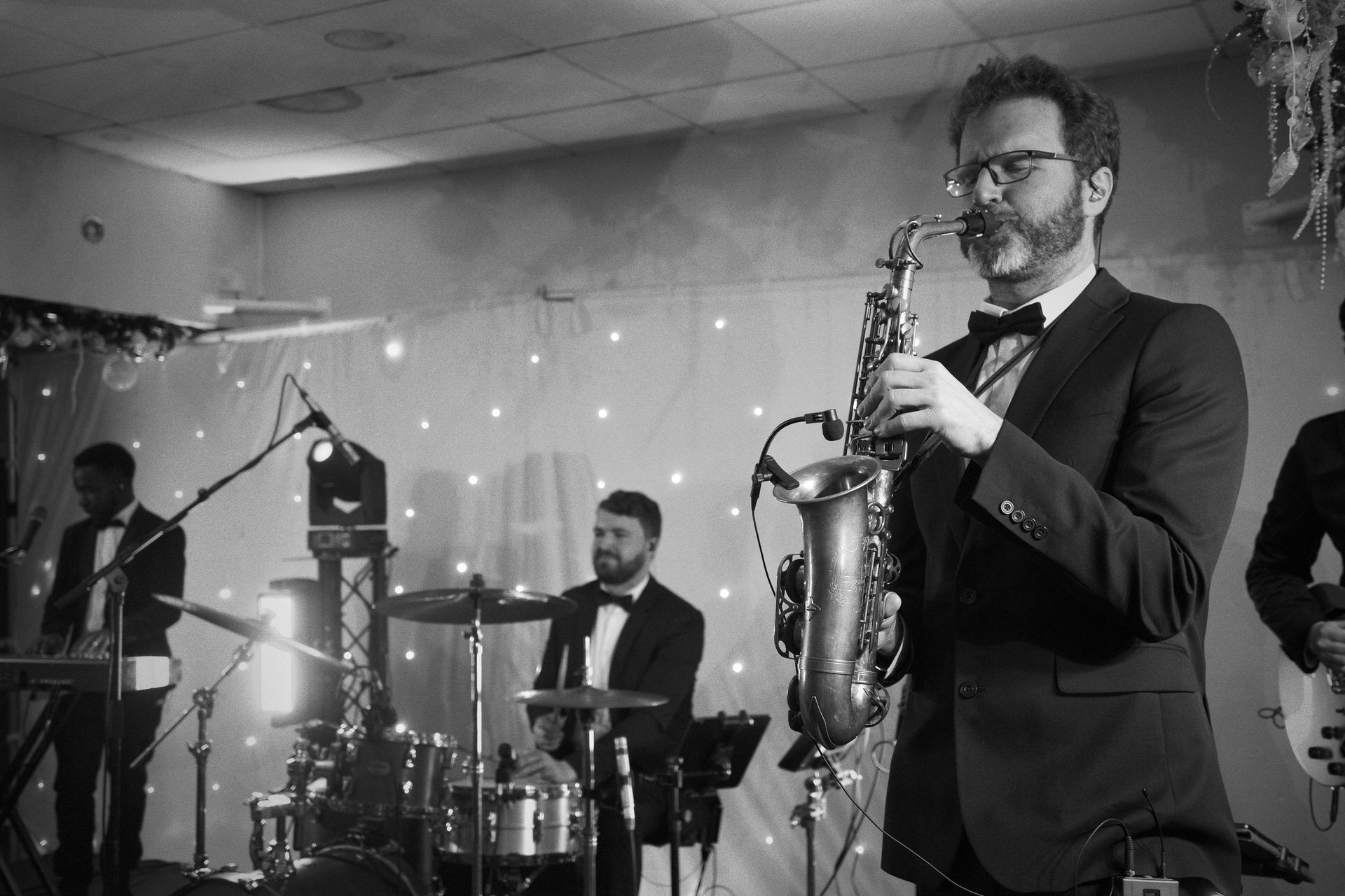 A tuxedoed saxophonist plays on stage while a drummer and keyboardist perform behind him, fairy lights glowing along the backdrop.