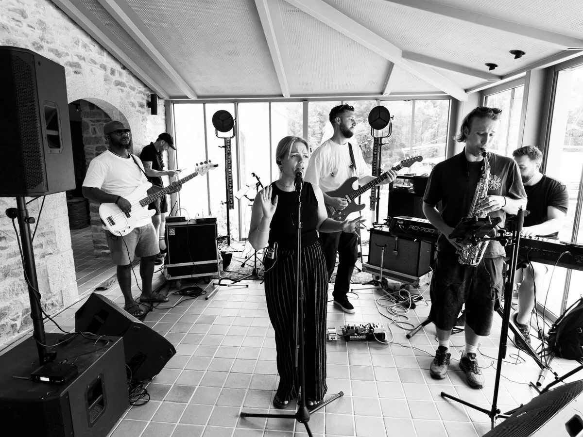 A live band performing in a sunroom, featuring a female lead singer, bassist, guitarist, saxophonist, and keyboardist.