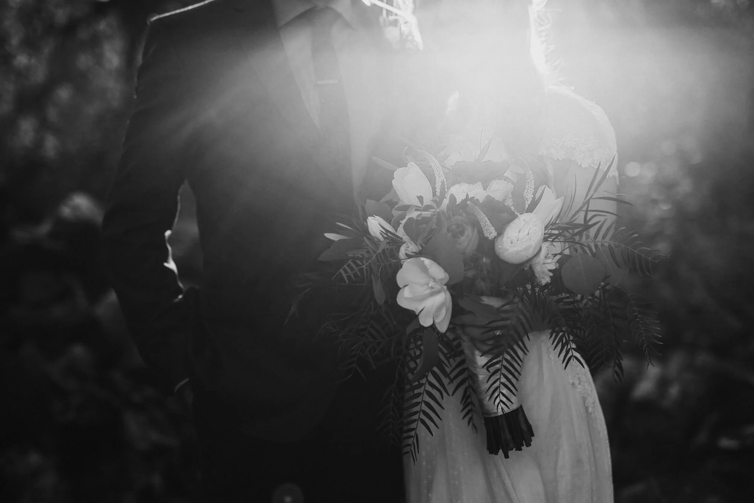 Bride and groom stand close outdoors, sun flaring behind them. She holds a lush bouquet of flowers, their faces partly hidden by bright light.