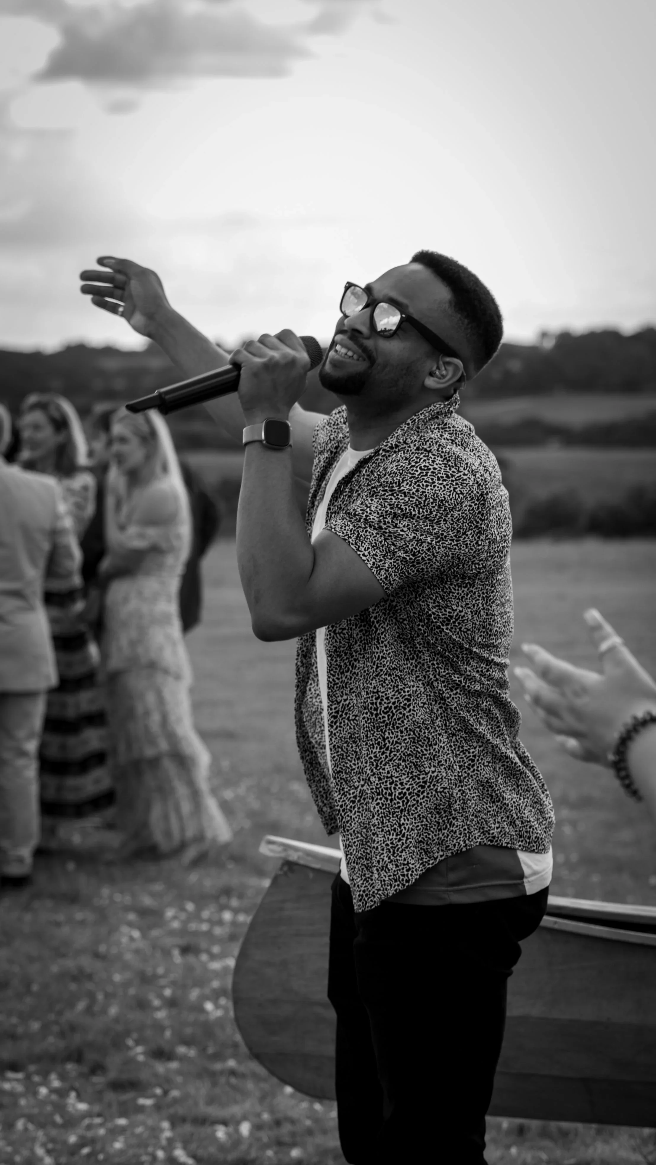 A smiling singer in sunglasses holds a mic outdoors, arm raised. Guests behind him clap and watch across an open field under a cloudy sky.