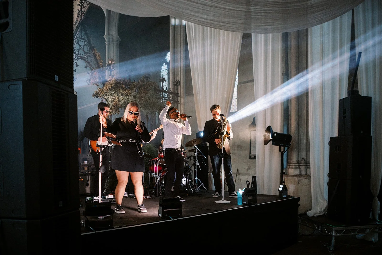 A live band performs on a small stage in a grand hall, bright beams of light cutting through white drapes as singers and musicians play energetically.