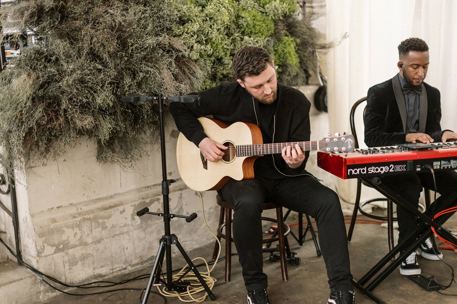 Two musicians sit indoors: one softly plays an acoustic guitar, the other a red keyboard, greenery hanging behind them.